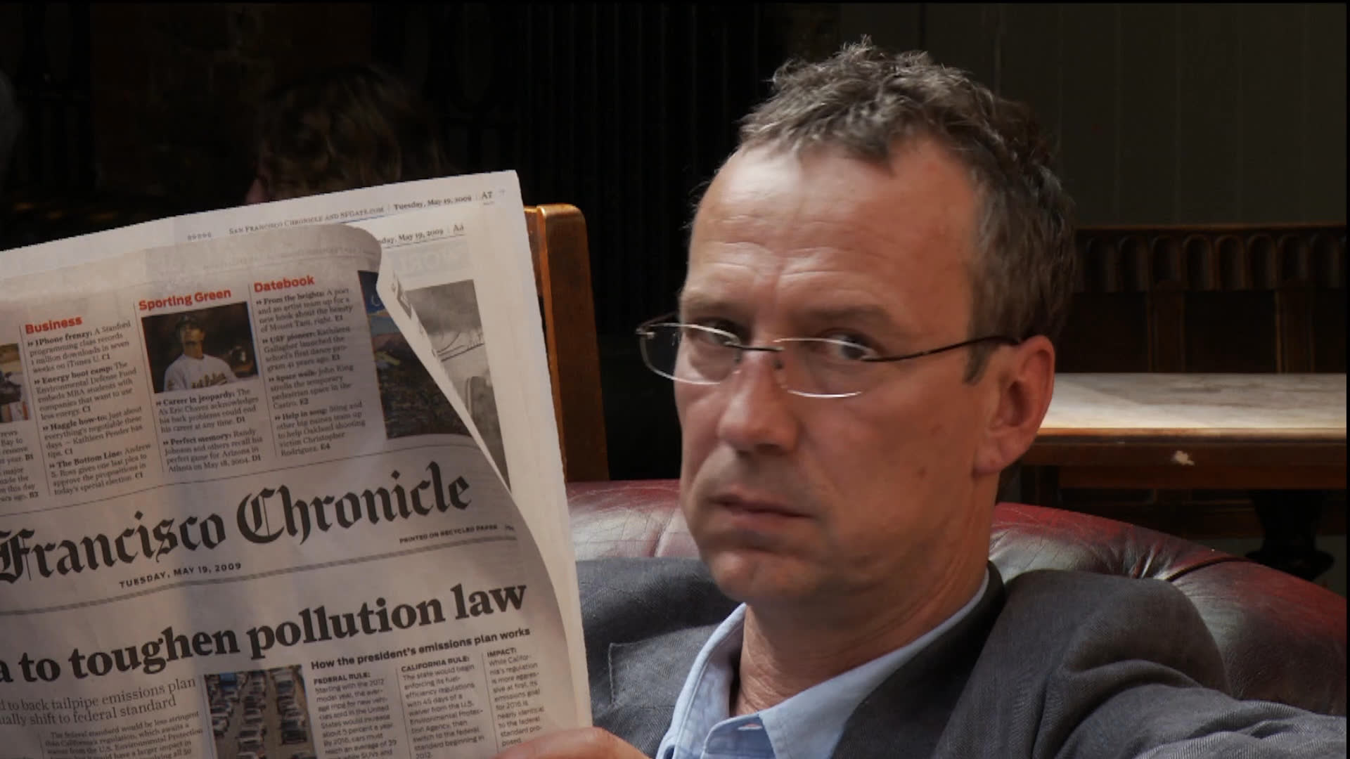 Two men sit together on a leather sofa, engaging in a focused conversation while intermittently reading from newspapers in a relaxed indoor setting.