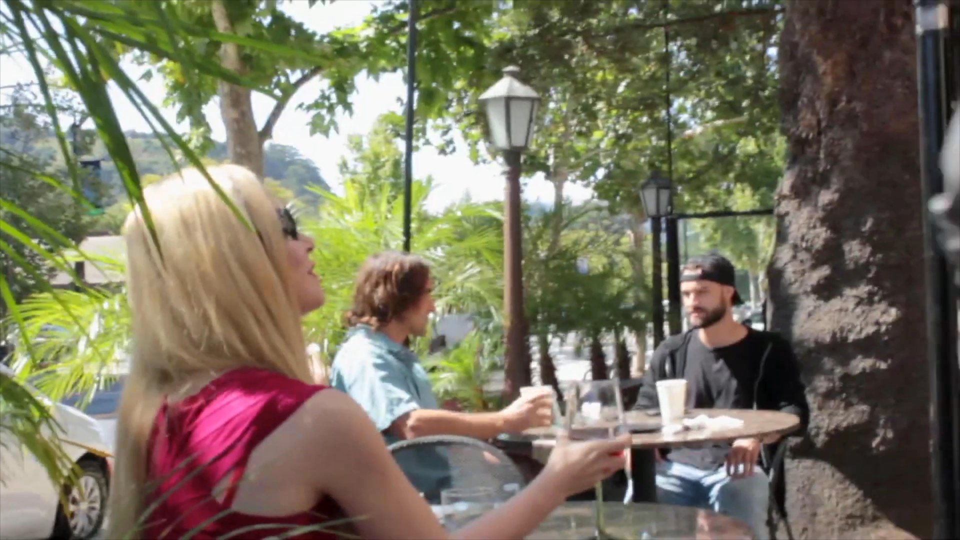 A woman in a red top enjoys a drink at an outdoor cafe before being joined by a man in a trench coat for a conversation. They engage in a serious, focused dialogue at their table.