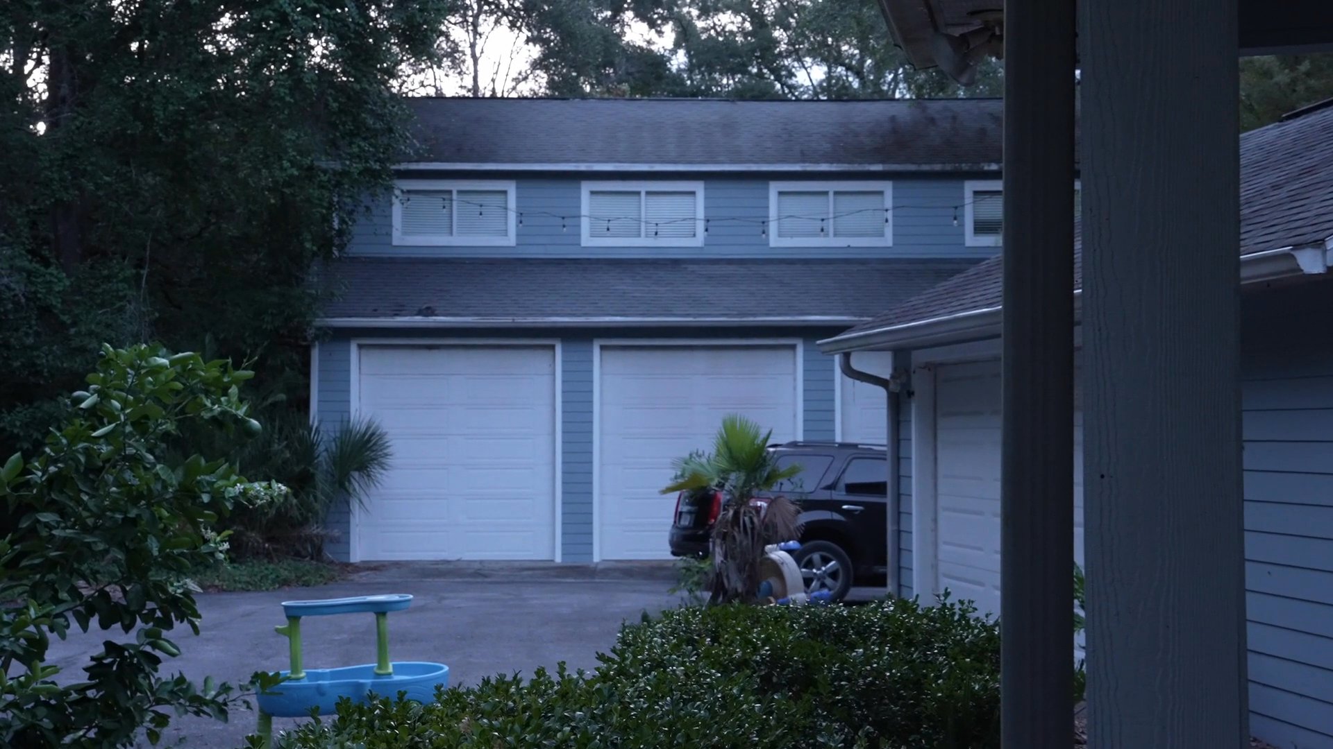 Individuals engage in conversation within a residential kitchen before the scene transitions to a view of the suburban exterior and a woman preparing food.