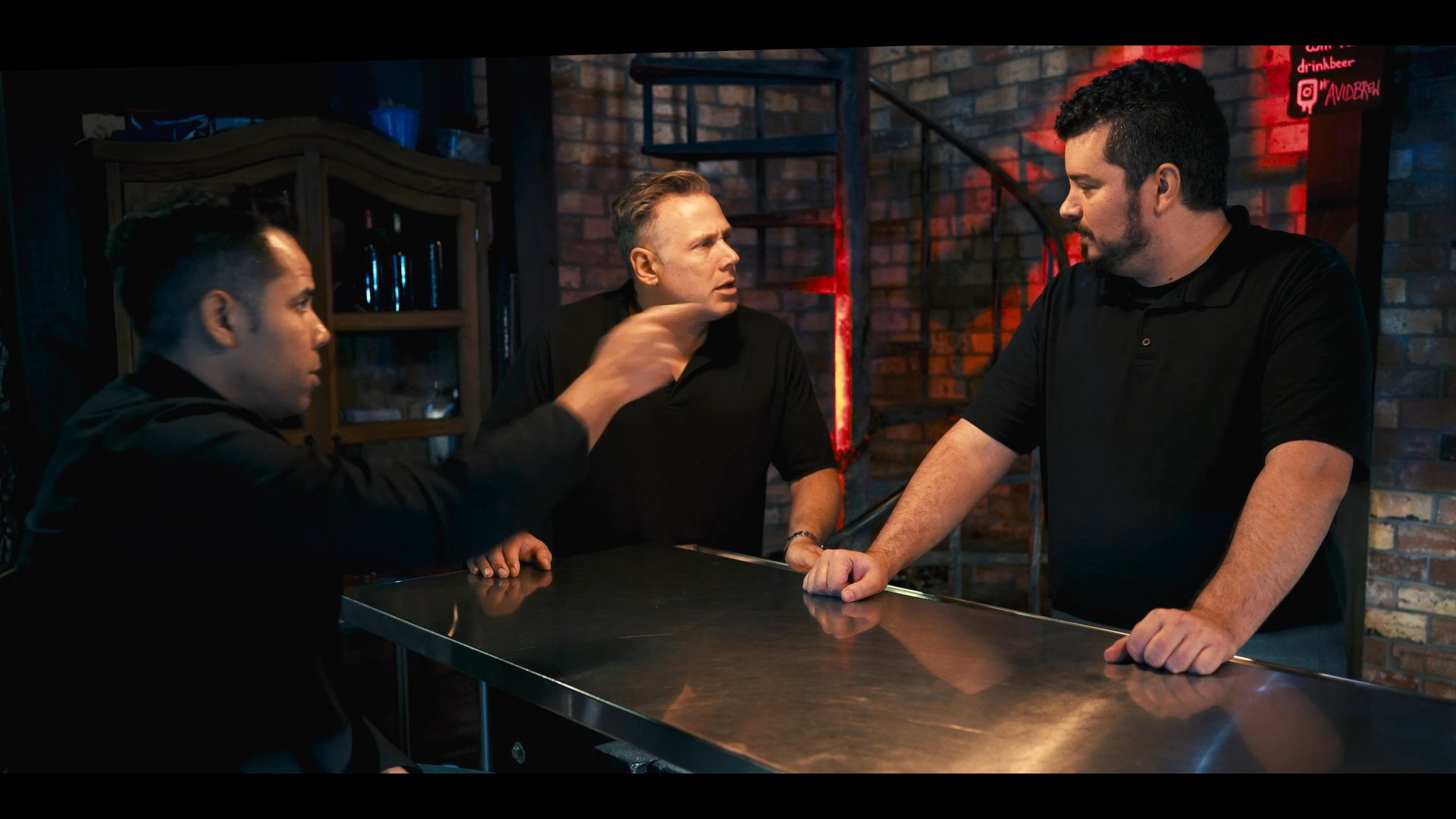 Three colleagues engage in a focused discussion at a bar counter, sharing ideas in a professional environment. They maintain active communication while leaning on the stainless steel surface.