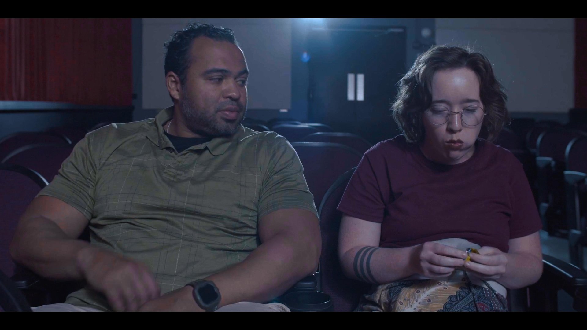 Two individuals sit side-by-side in a dimly lit cinema, sharing snacks and engaging in a casual conversation while waiting for a film.