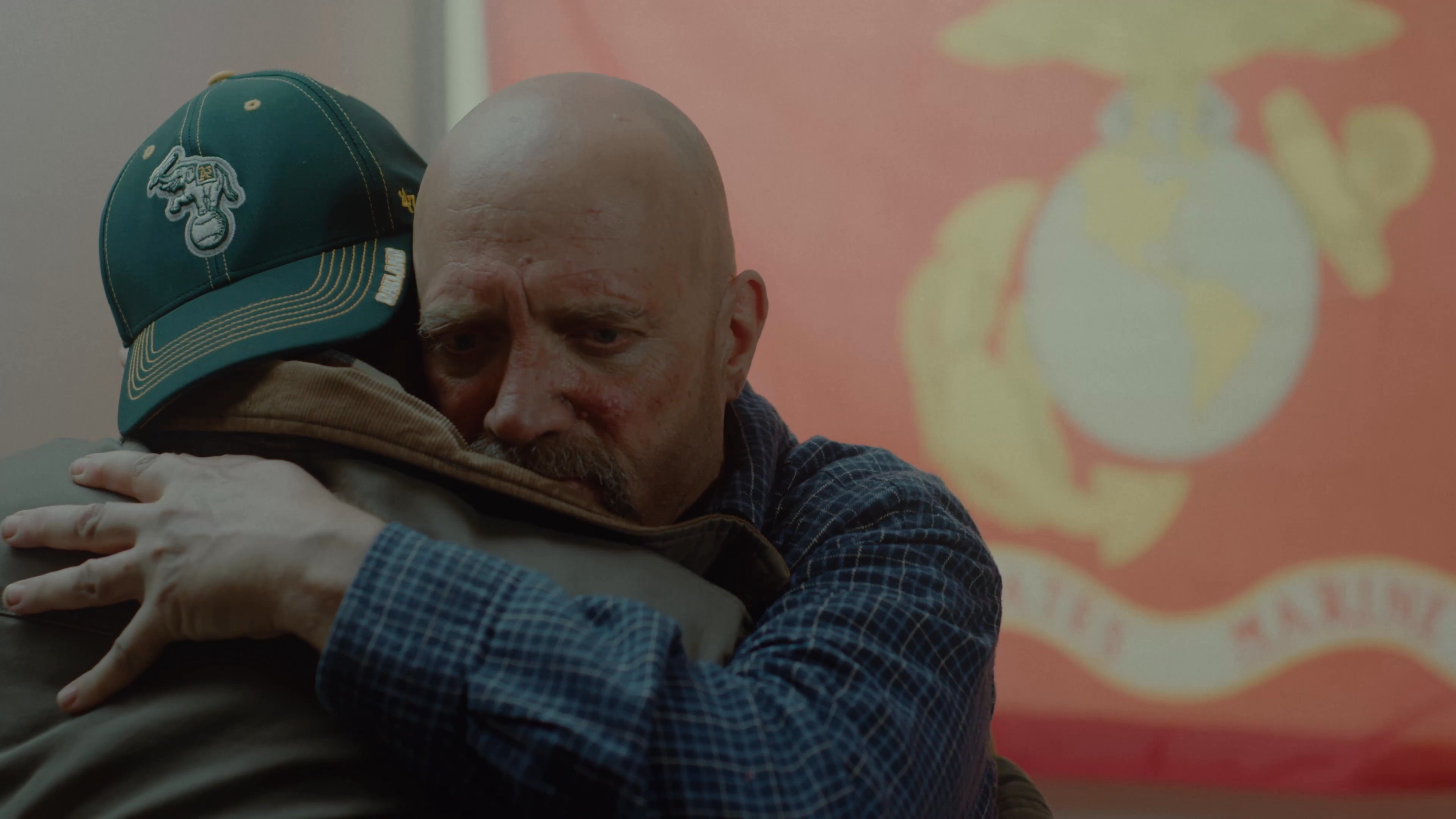 A man returns home and shares an emotional embrace with an older man sitting in a living room decorated with military memorabilia. The two share a tender, heartfelt moment of connection and comfort.
