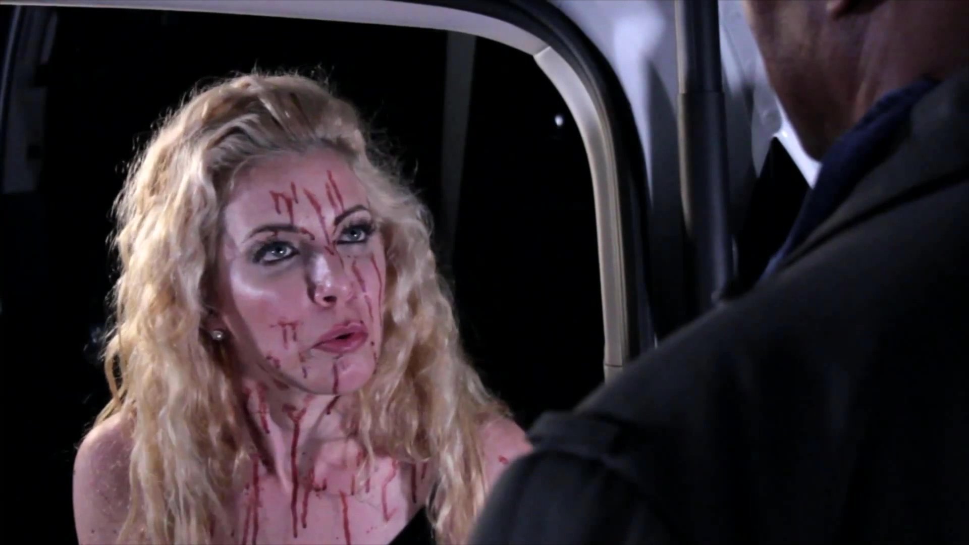 A woman with blood on her face engages in an intense verbal confrontation with a man inside a dimly lit space. The interaction is charged with palpable tension as they exchange dialogue.