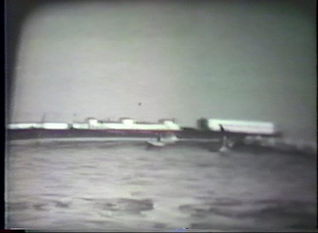 A surfer rides a wave along a coastal pier in a grainy, black-and-white archival sequence. The footage captures the rhythmic motion of the surfer maintaining balance amidst the breaking surf.