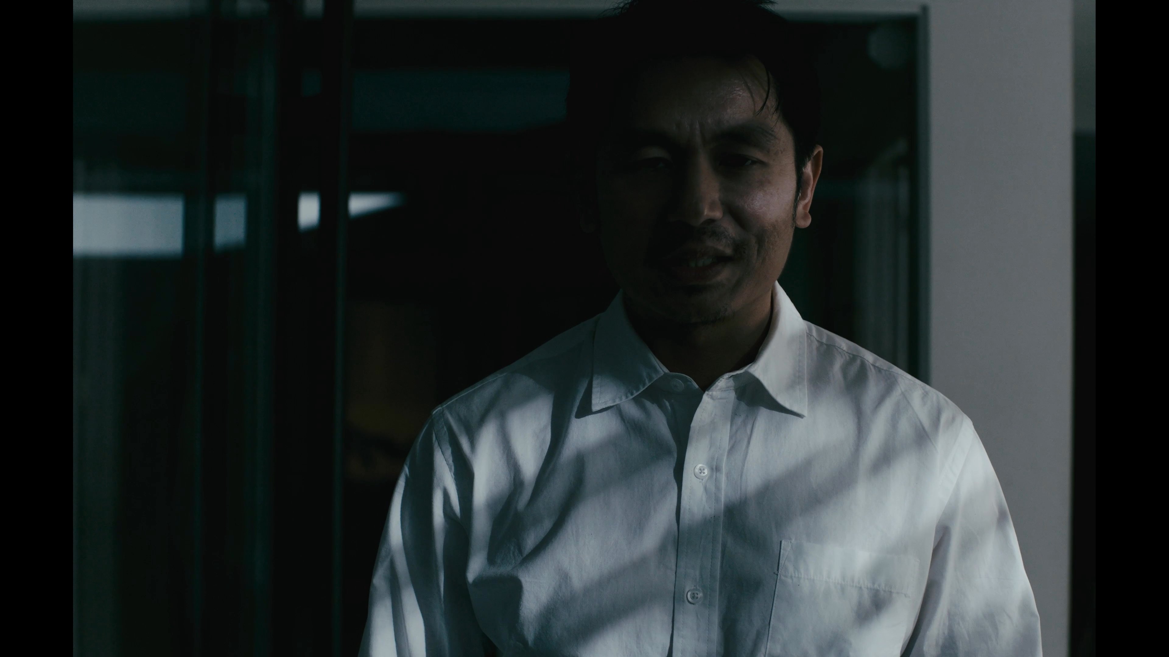 A man in a white shirt enters a modern home, where a woman awaits him. They share a quiet moment of acknowledgement in the dimly lit interior.