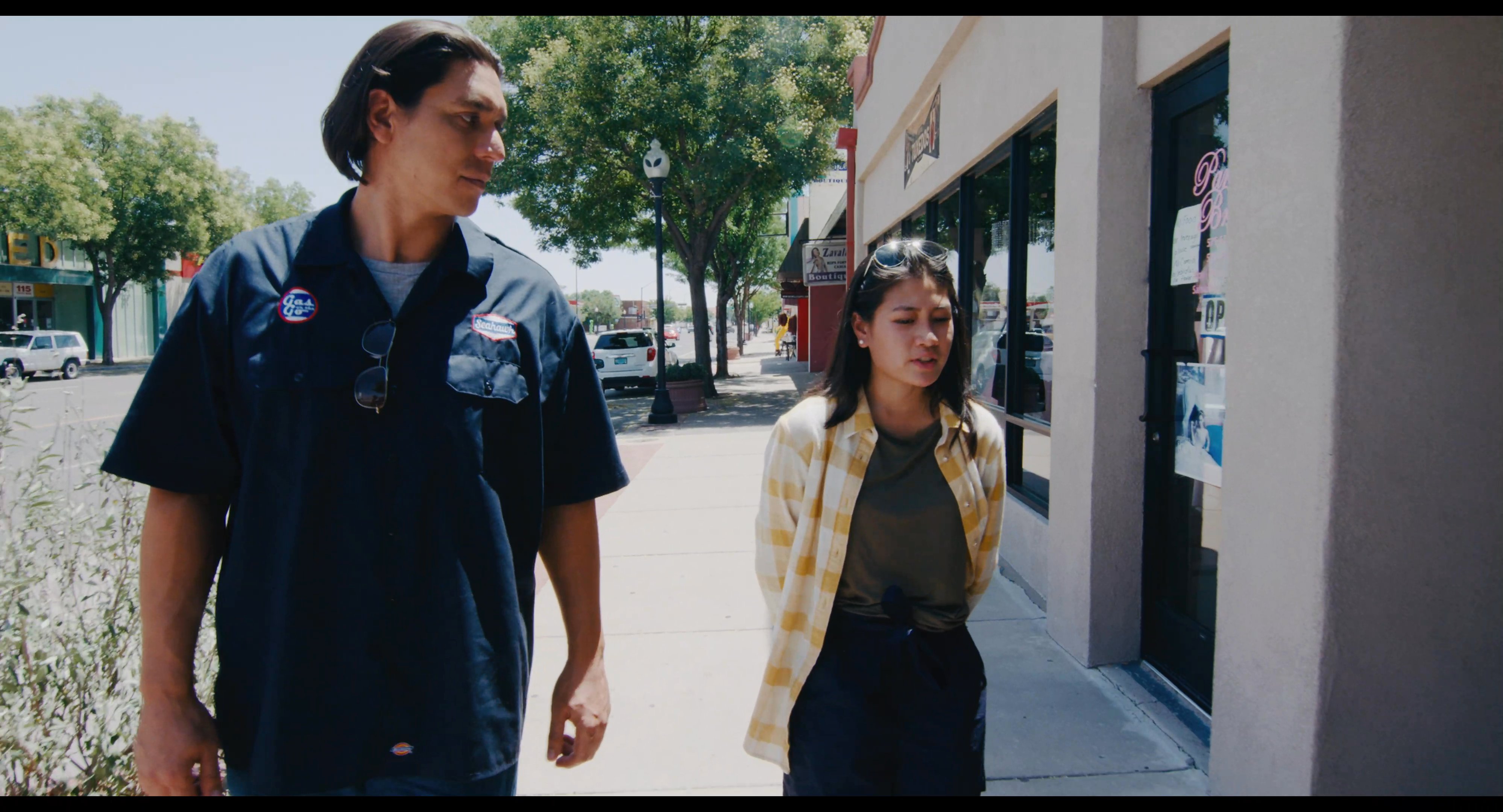 A man and a woman walk together along an urban sidewalk past storefronts on a bright, sunny day. The scene shifts to a wide angle view showing the street corner and local business architecture.