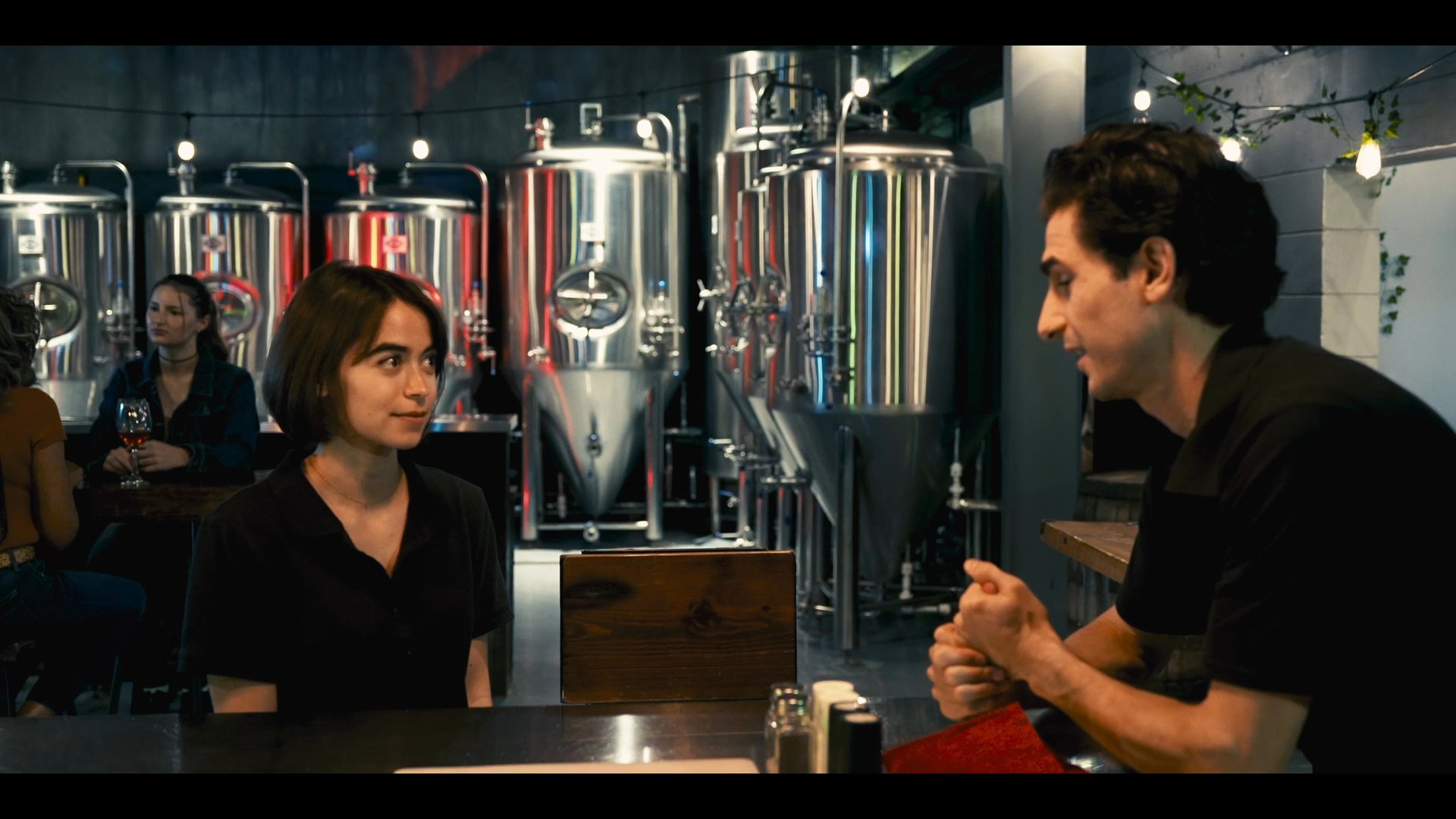 Individuals engage in conversation within a brewery setting, with metal brewing vats visible in the background. A woman checks her phone while seated at the bar, reflecting a casual social environment.