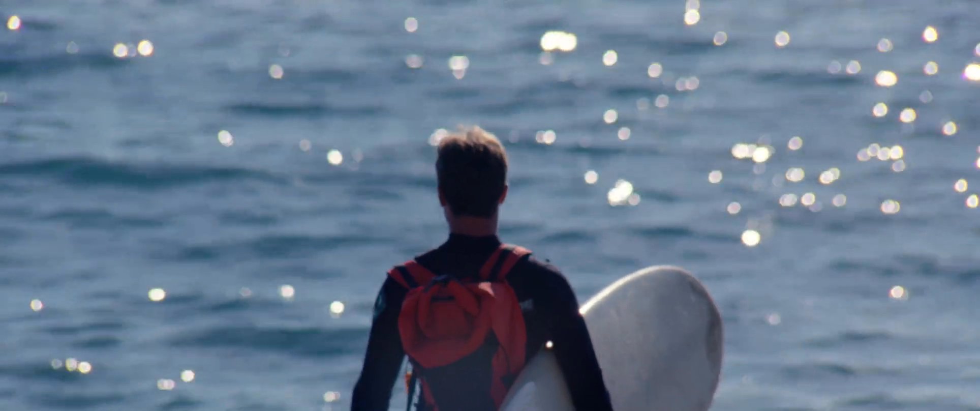 A surfer dons a wetsuit and carries a surfboard toward the ocean along a sunlit coastal beach.