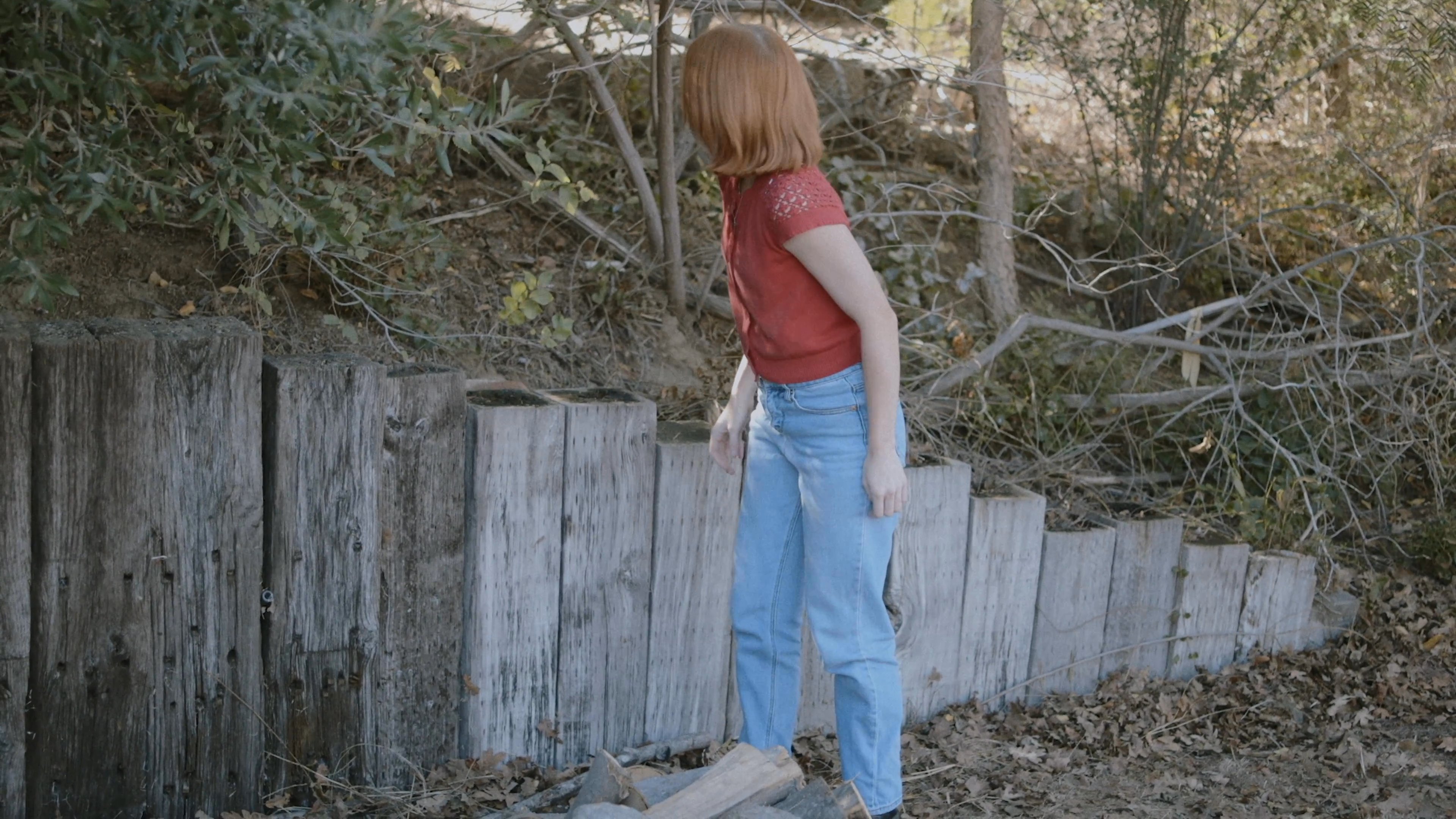 A woman stacking firewood in a wooded area abruptly pauses, appearing alert and unsettled before abandoning her task to retreat into the forest.