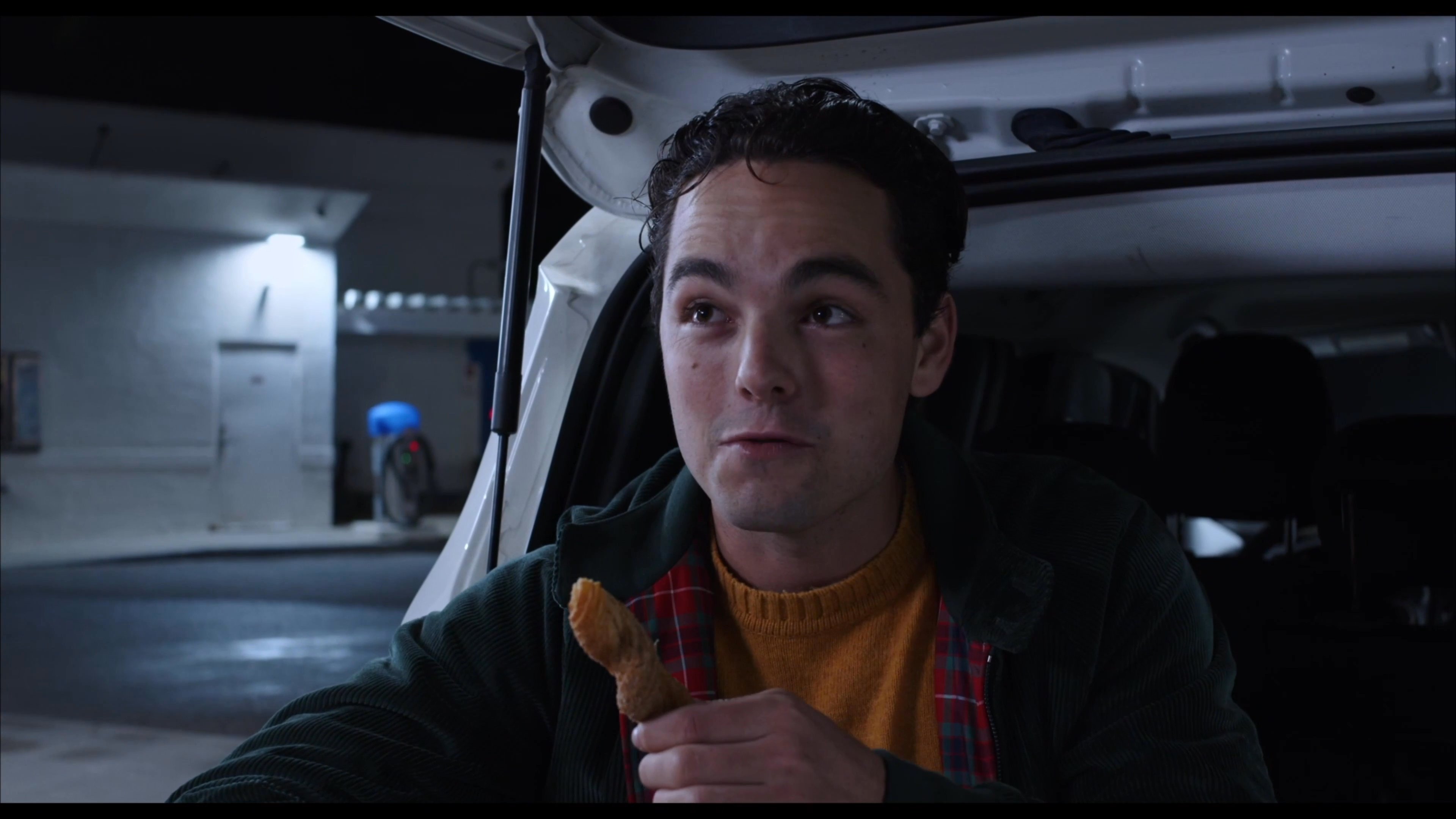 Two men engage in a casual conversation at a gas station during the night, with one individual eating a snack while talking.