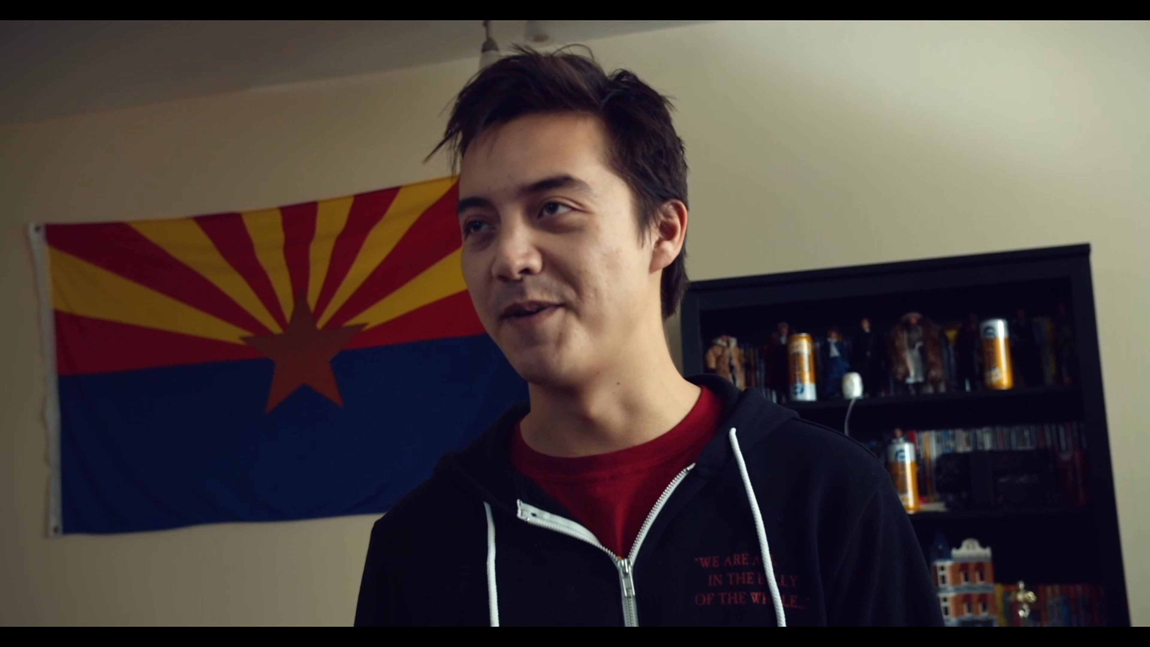 Two young men engage in an informal conversation within a decorated living space featuring an Arizona state flag. The scene shifts between their dialogue and close-up views of personal photographs pinned to a wall.