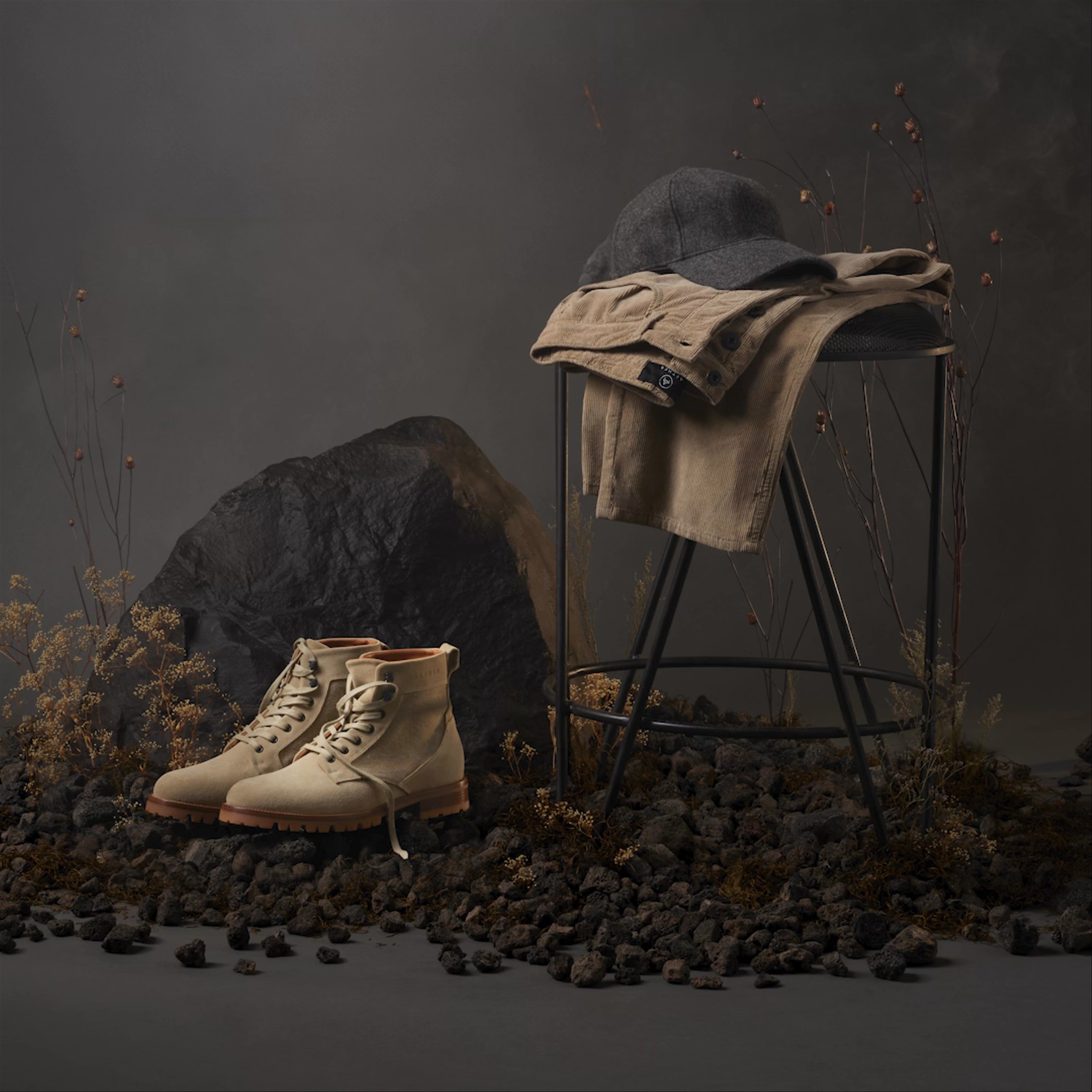 Studio scene on black background of men's and women's clothing on vintage stool alongside boots sitting amongst lava rocks and foliage