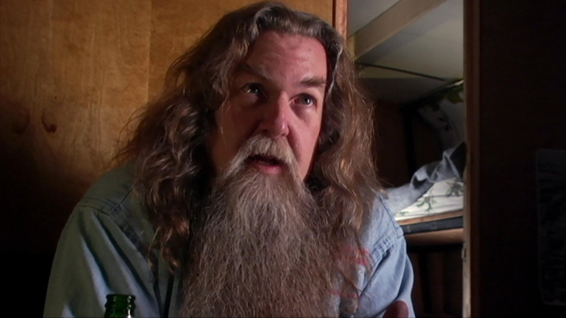 An aging musician with a long beard discusses his musical history while archival footage displays a past performance of him playing guitar alongside a violinist.