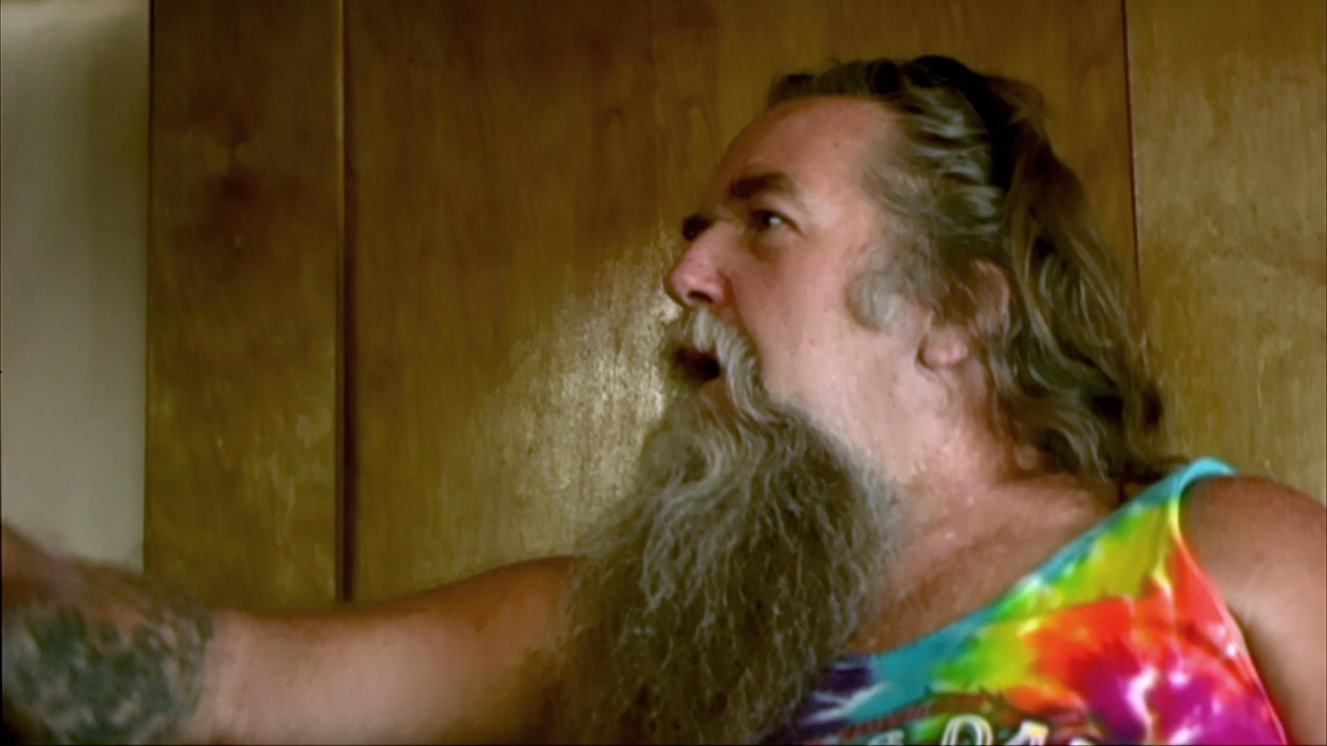 A long-haired, bearded man wearing a tie-dye tank top discusses his experiences while driving along a remote highway. The scene alternates between his perspective from the driver's seat and an intimate conversation inside the vehicle's cabin.