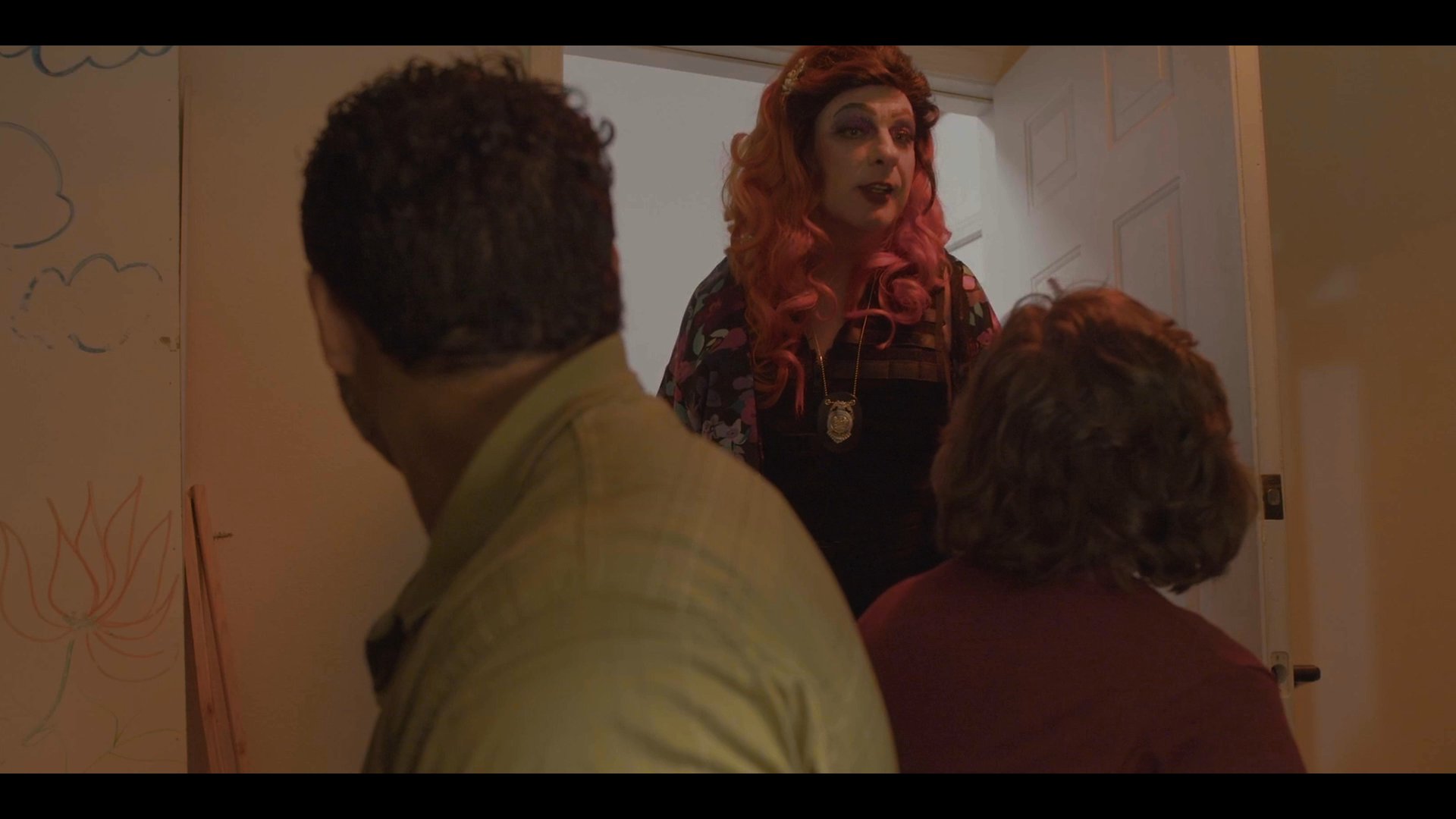 Two individuals engage in a tense conversation in a room before a third person in drag appears at the doorway, prompting further dialogue.