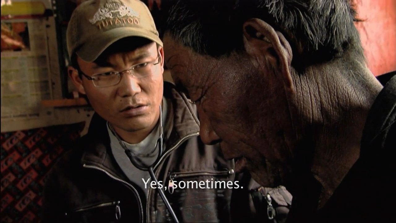 A physician conducts a physical examination of a patient in a rural setting, using a stethoscope to assess his condition while discussing symptoms. The interaction takes place in an austere interior space, highlighting the delivery of healthcare in remote regions.