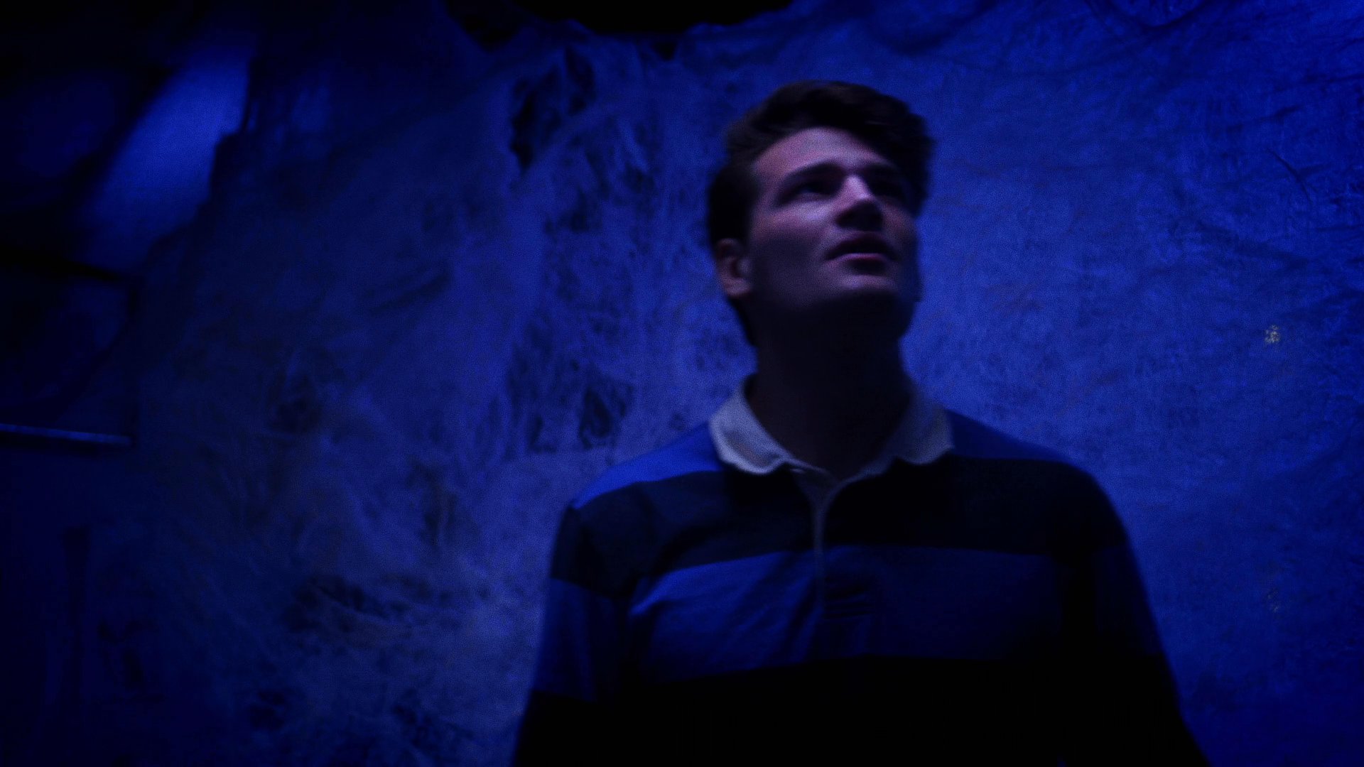 A man in a blue-striped shirt cautiously enters a dimly lit room covered in thick cobwebs. He observes the unsettling environment with apprehension as another figure appears in the background.