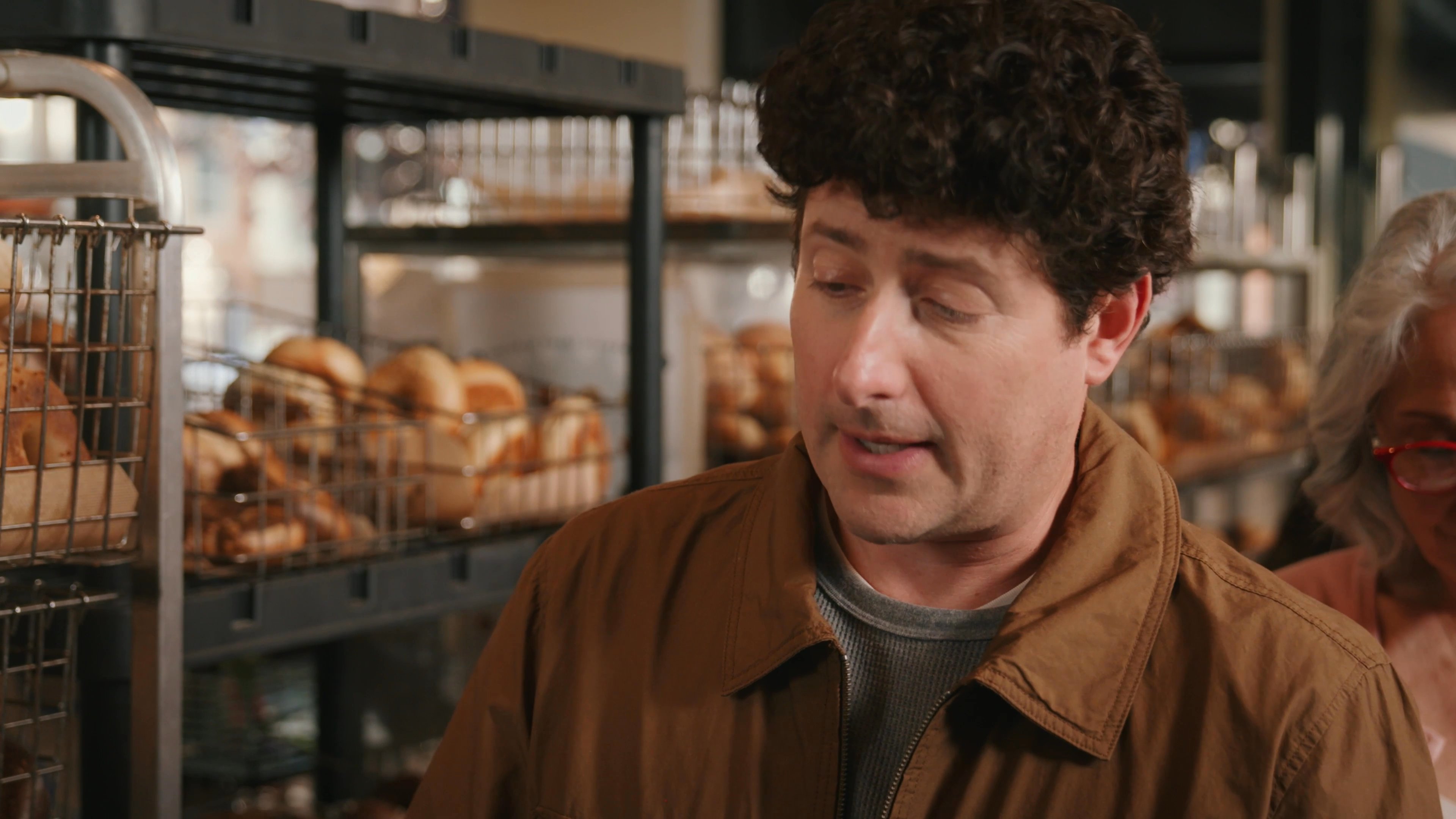 A man and an older woman engage in a conversation while standing near display racks in a bakery. The interaction captures a polite exchange between customers in a retail environment.