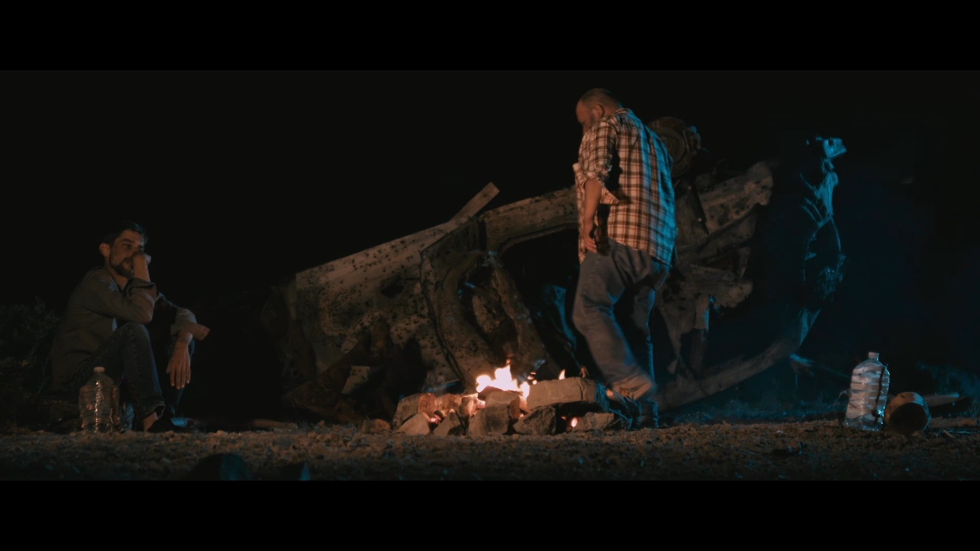 Two men sit by a campfire at night, where one individual retrieves an object from near a vehicle wreck and presents it to the other.