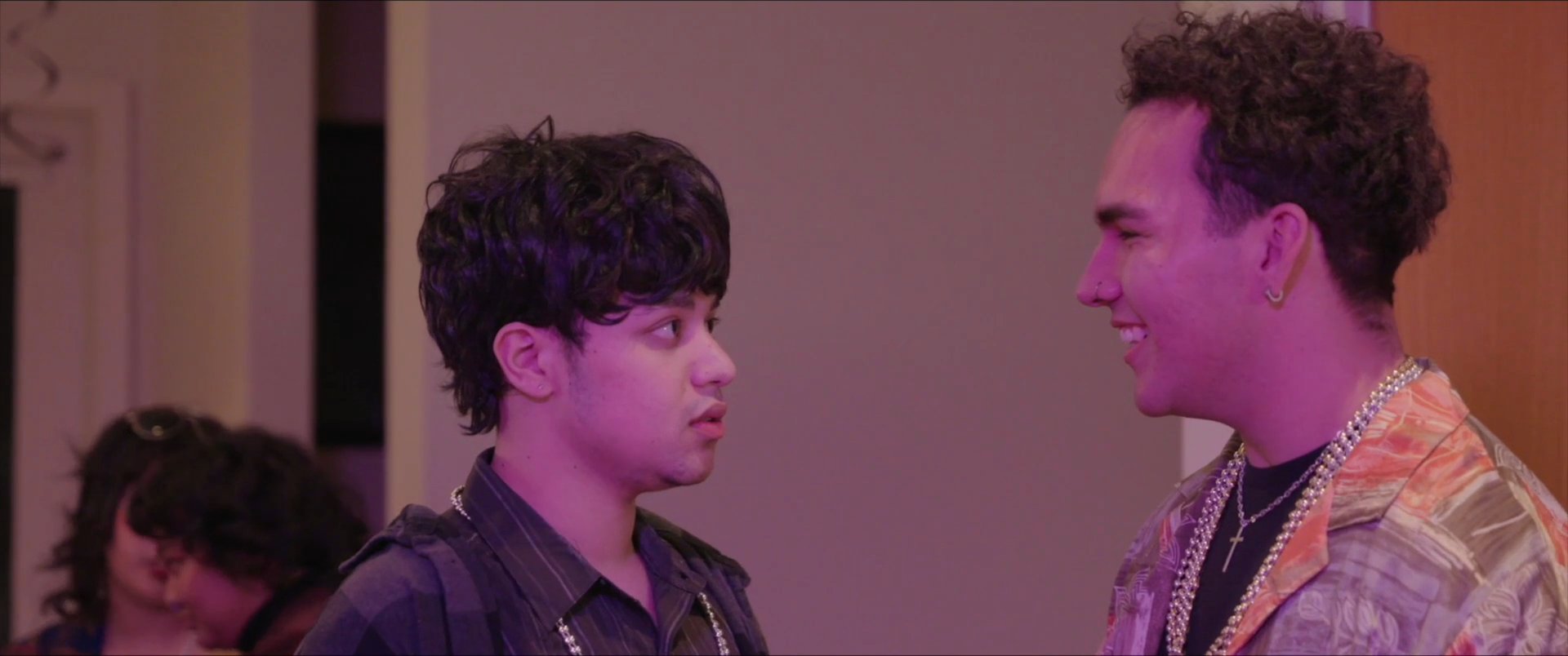 Two young men engage in a private, focused conversation amidst a lively indoor social gathering. The interaction concludes with a transition to an exterior nighttime setting.