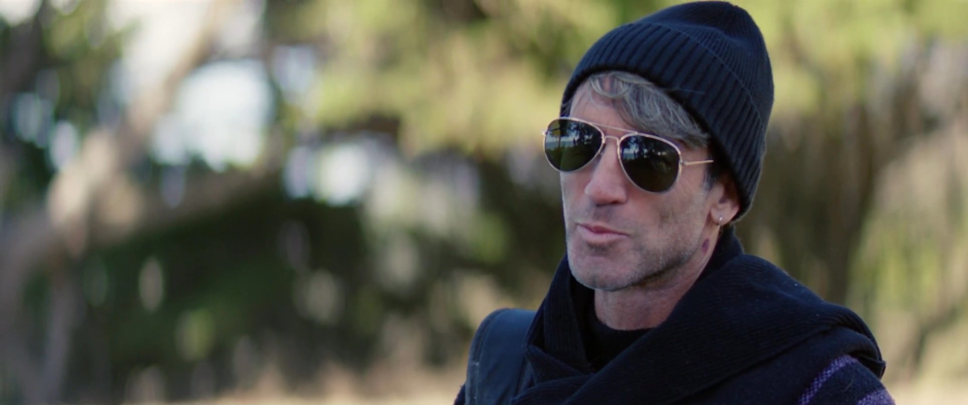 Two men engage in an outdoor conversation near the coast, with one individual wearing sunglasses and a beanie while the other listens attentively.