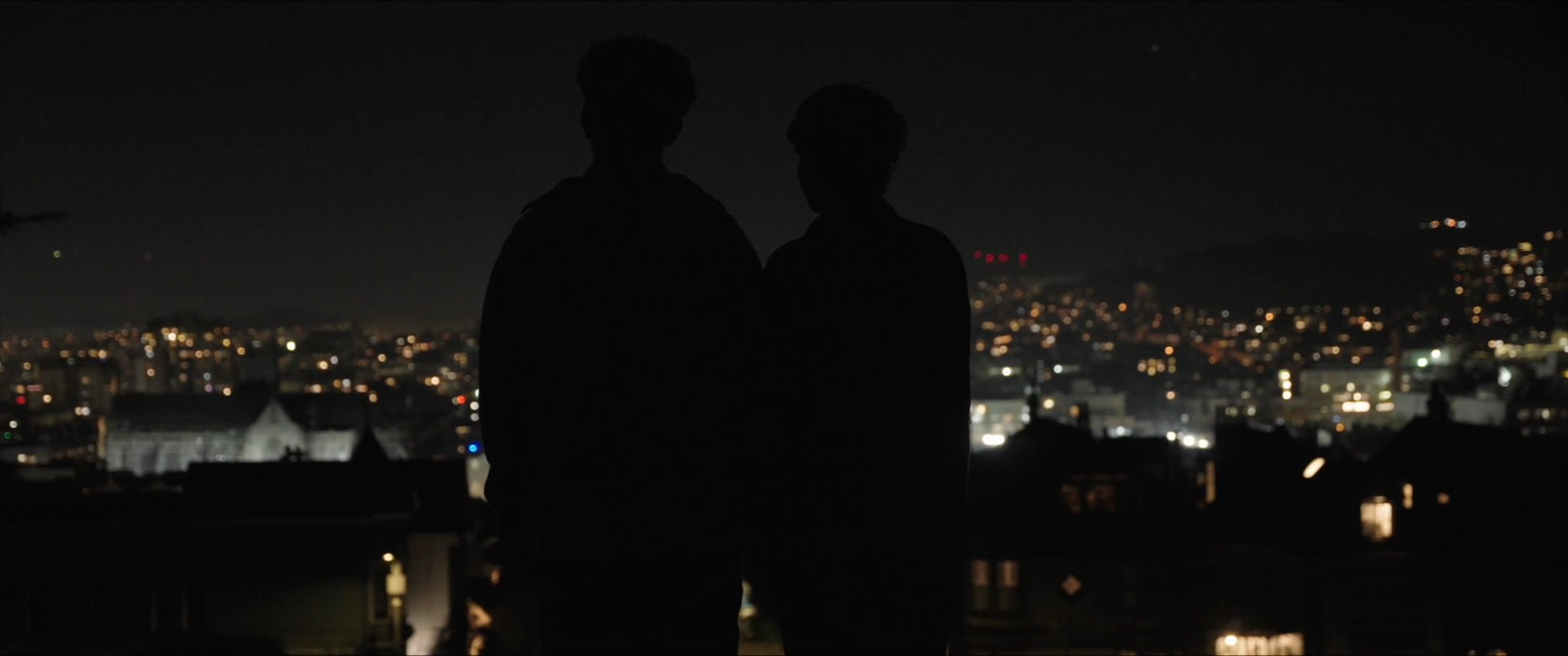 Two individuals share an intimate conversation overlooking a glowing cityscape at night, which transitions into a formal scene featuring a group of men gathered at a podium.