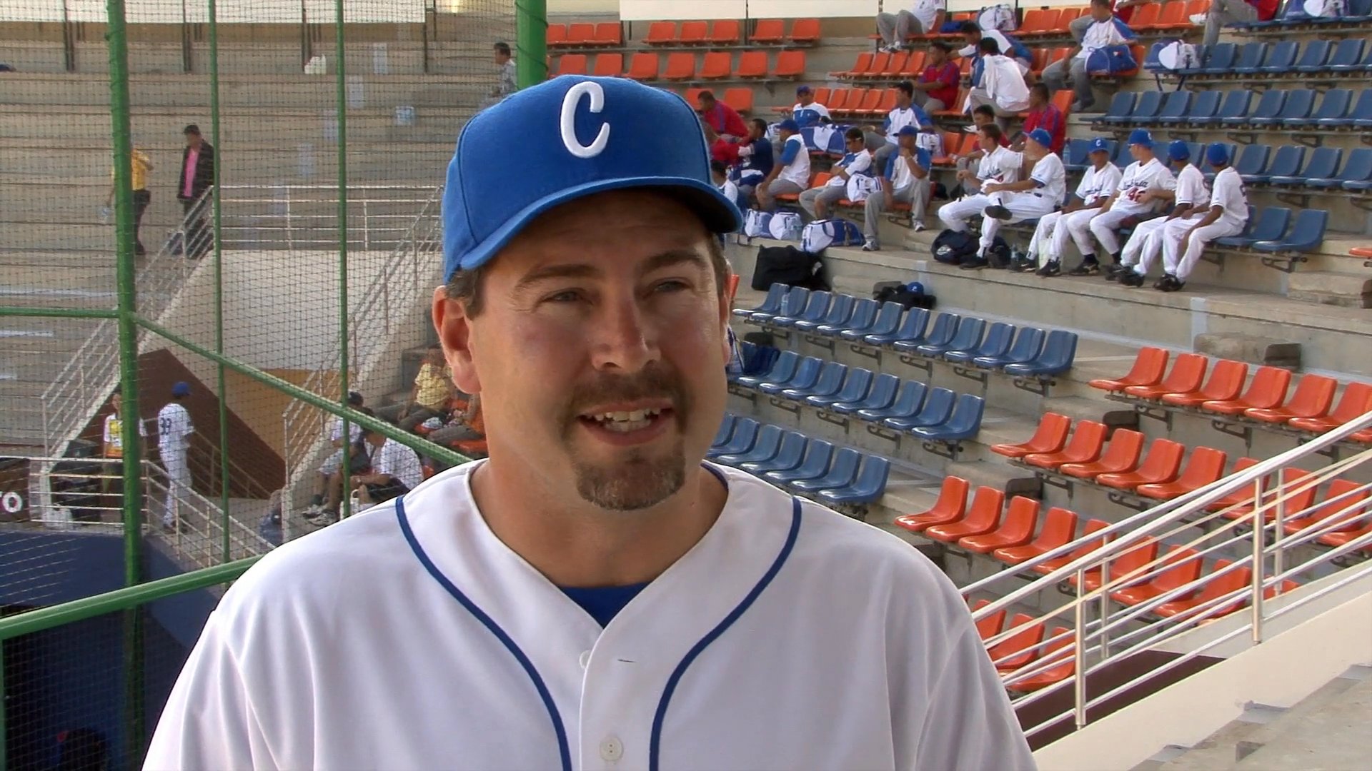 Two individuals in baseball uniforms provide commentary while standing in front of stadium bleachers. The scene transitions between a coach and a player sharing insights from the field.