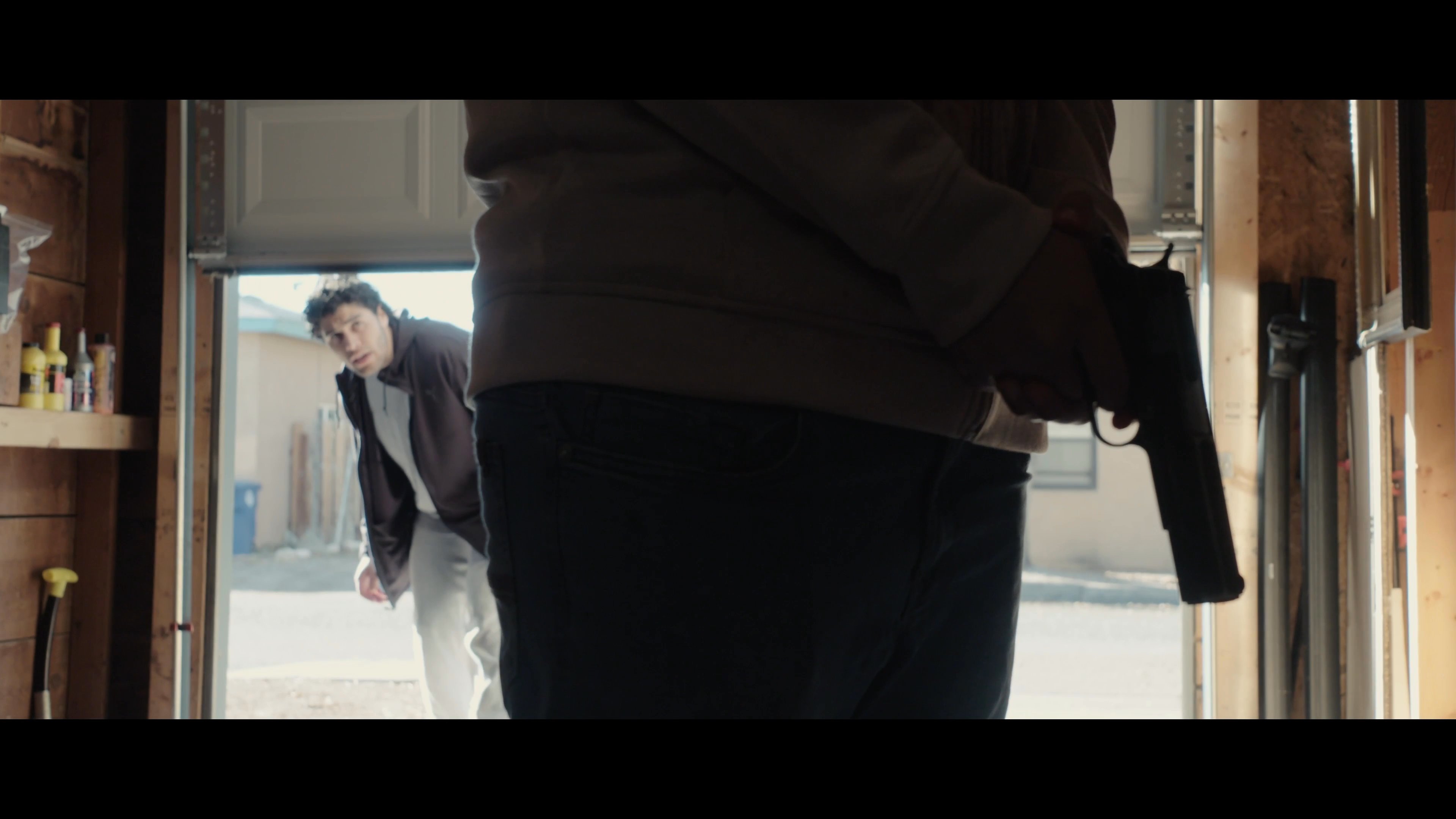 Two women confront a man covered in bloodstains inside a garage, leading to a tense exchange before the man retreats to handle a firearm. The scene captures an atmosphere of high emotional distress and unresolved conflict.
