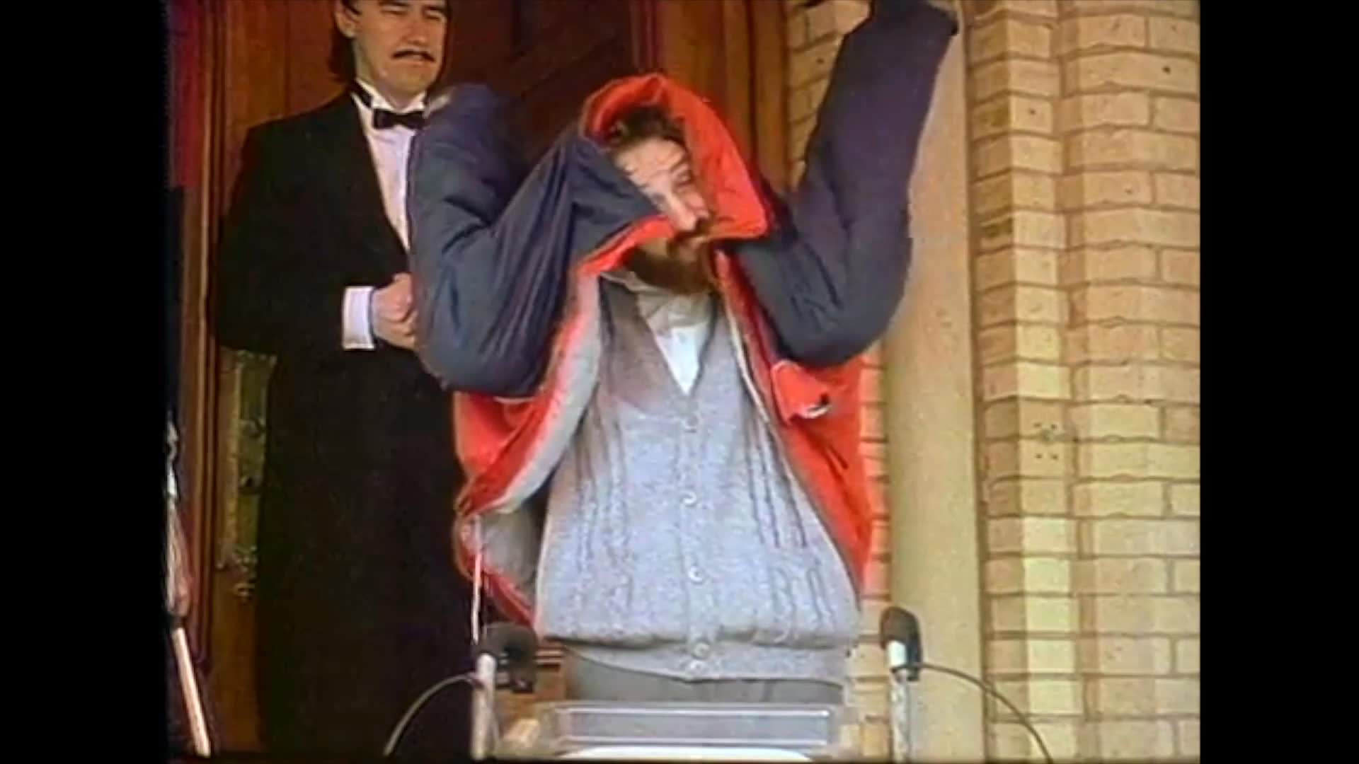 A formal butler assists a bearded gentleman by helping him put on a winter jacket outside a residential entrance.