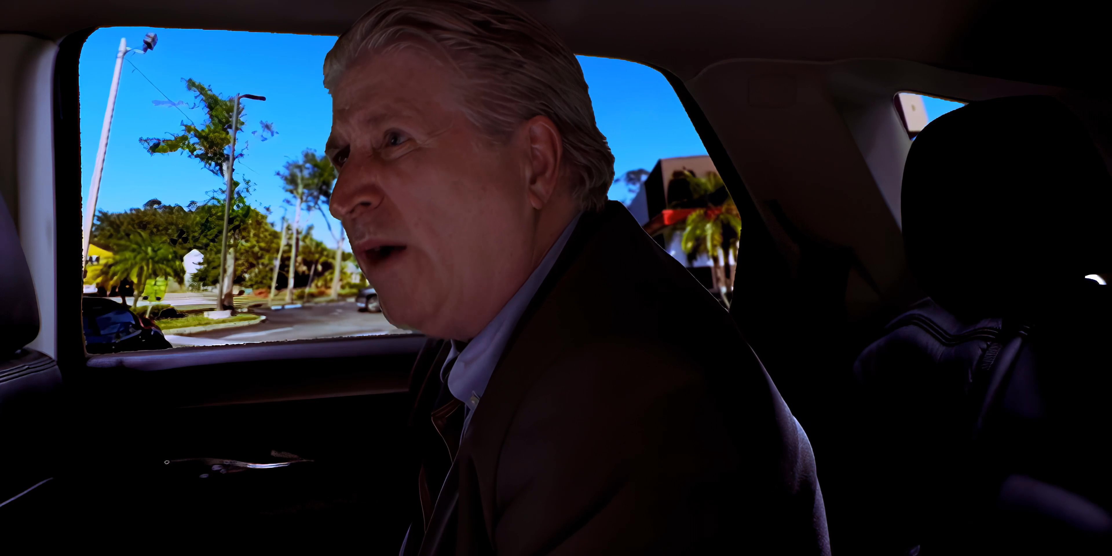 Two men discuss topics while traveling in a car through an urban area, transitioning to a wide aerial view of a sprawling metropolitan cityscape near a harbor.