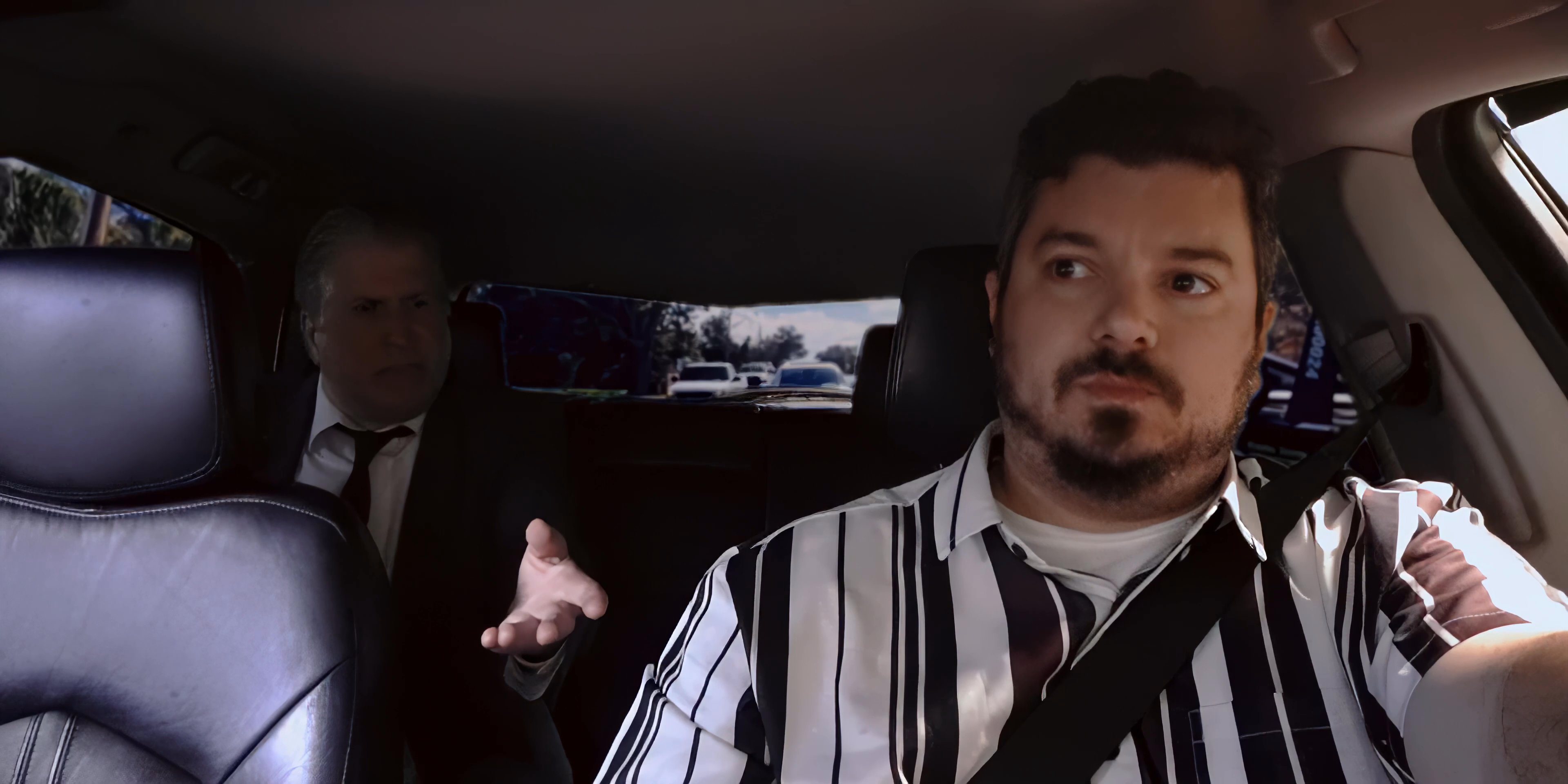 A rideshare driver and his passenger engage in a spirited conversation while traveling through a suburban neighborhood. The driver pauses to drink from a brown paper bag during the commute.