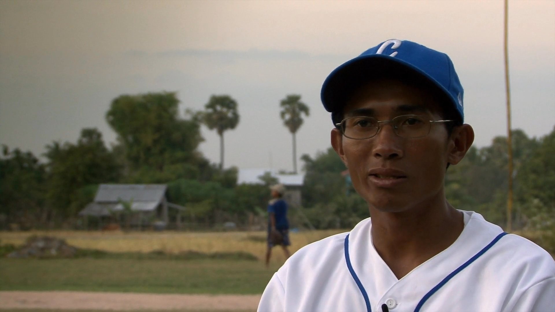 A man in a baseball uniform discusses his community experiences while standing on a rural field. The segment transitions to show local agricultural surroundings and people working near traditional shelters.
