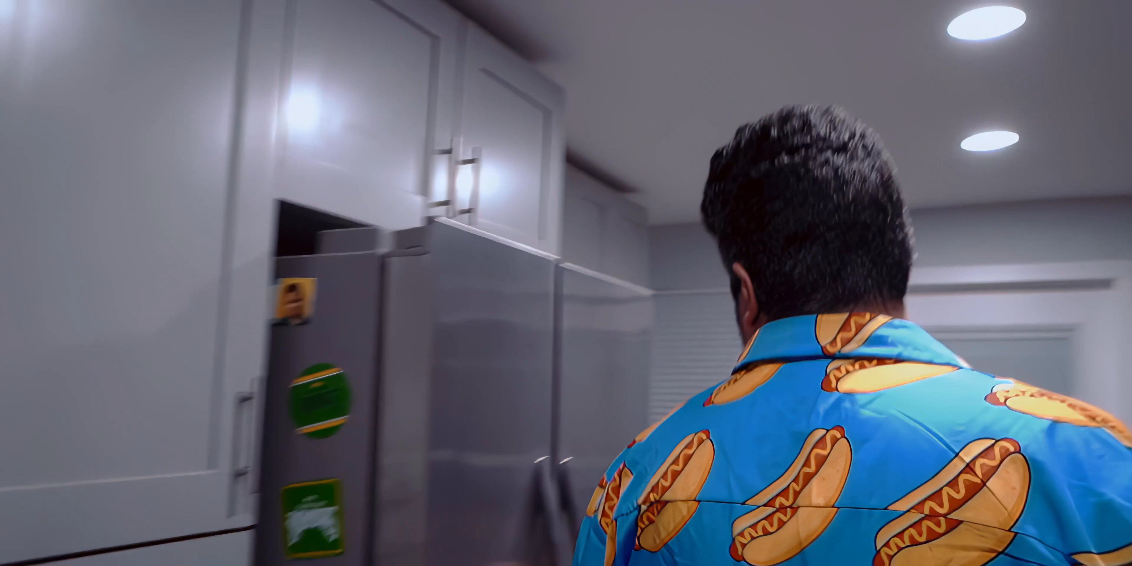 A man leaves a dark room and walks into a brightly lit kitchen to inspect the contents of his refrigerator during the night. He appears exhausted and hesitant as he looks for food.