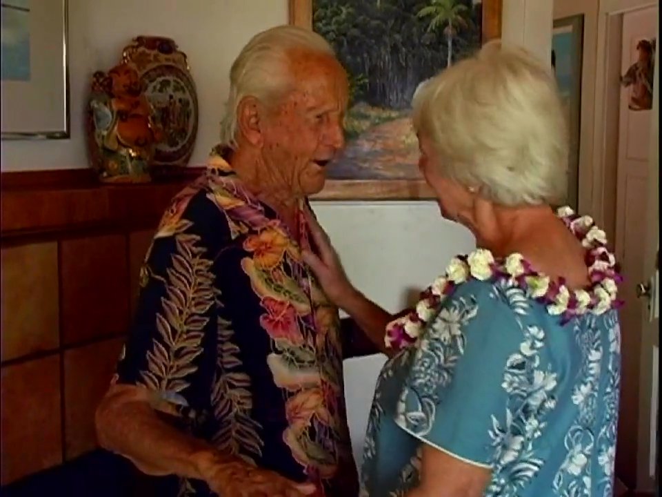 An elderly man and woman, both adorned with floral leis, exchange an affectionate, long-awaited embrace in a warm home environment. Their interaction captures a moment of deep personal connection and joy.