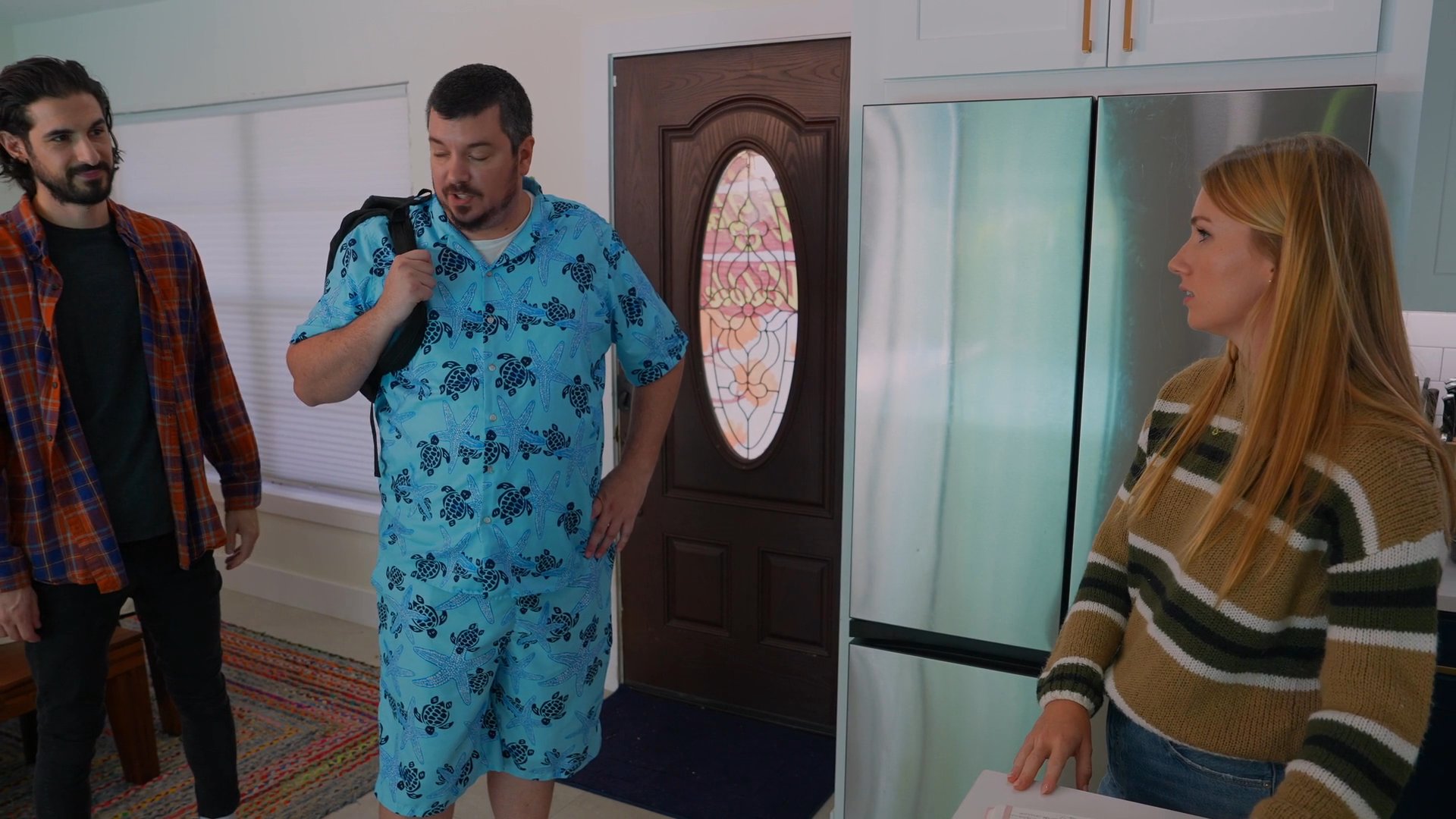 Two men enter a residential home to greet a group of people already present in the kitchen area. The arrival prompts a casual exchange and immediate social engagement between the guests and the residents.