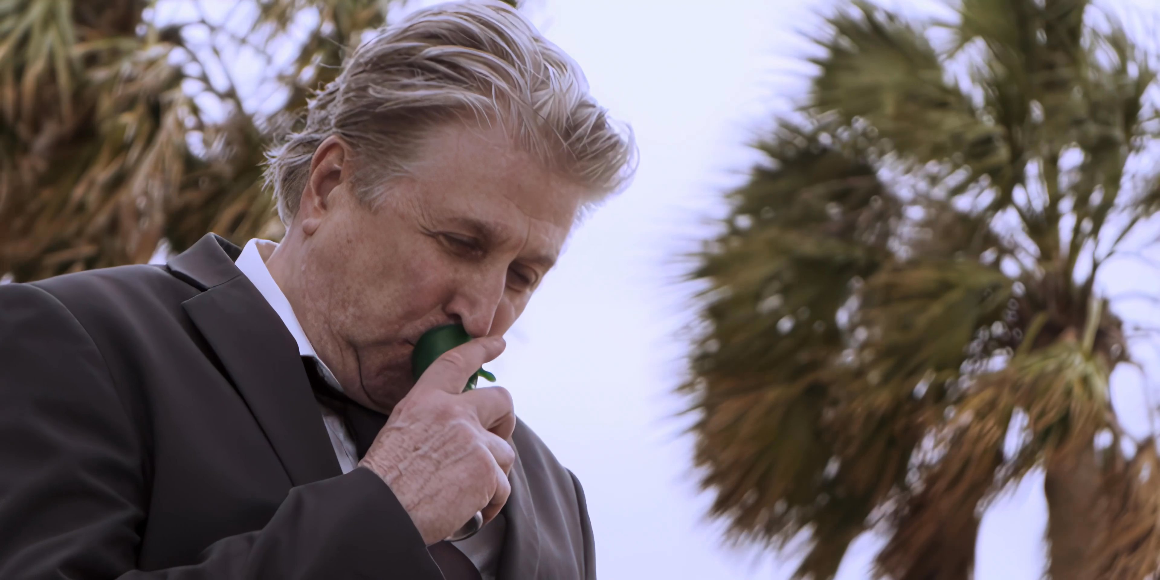 A man in a suit experiences physical distress and uses an inhaler while standing among palm trees. The scene shifts to reveal another man observing the situation.