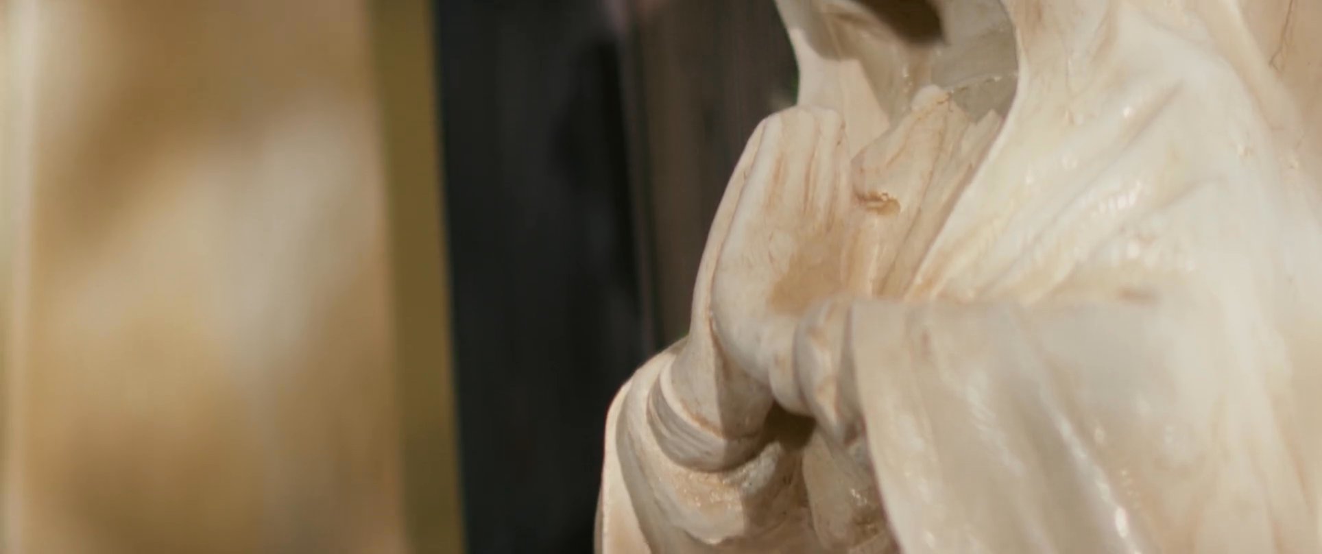 A hooded figure walks through a barren desert landscape, transitioning to intimate shots of religious statues, iconography, and traditional decor, evoking a sense of spiritual reverence.