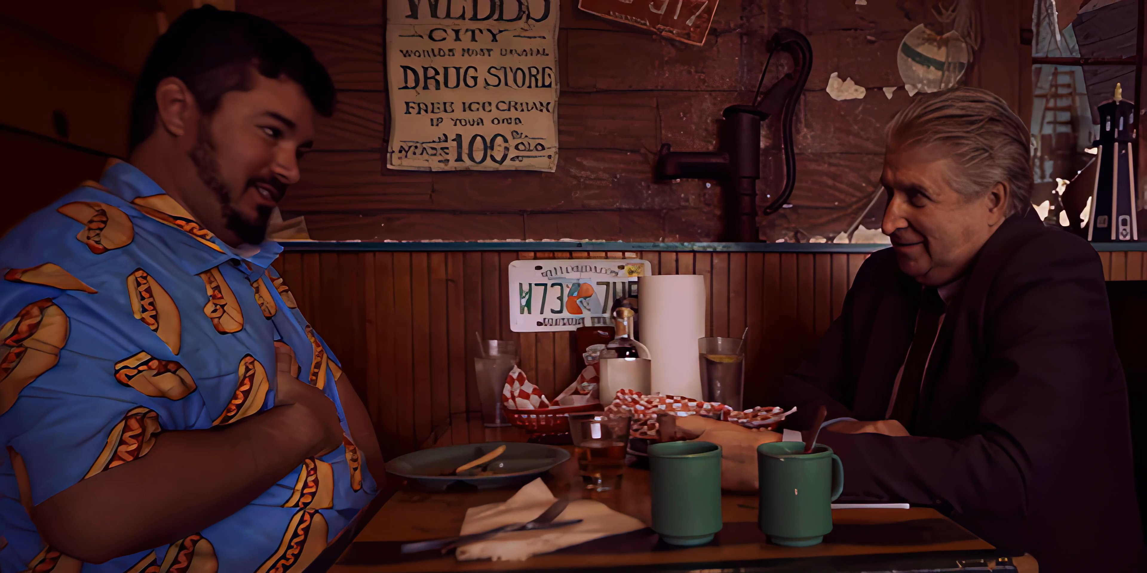 Two men engage in a focused conversation at a diner table before the scene transitions to an evening view of a lit city skyline.
