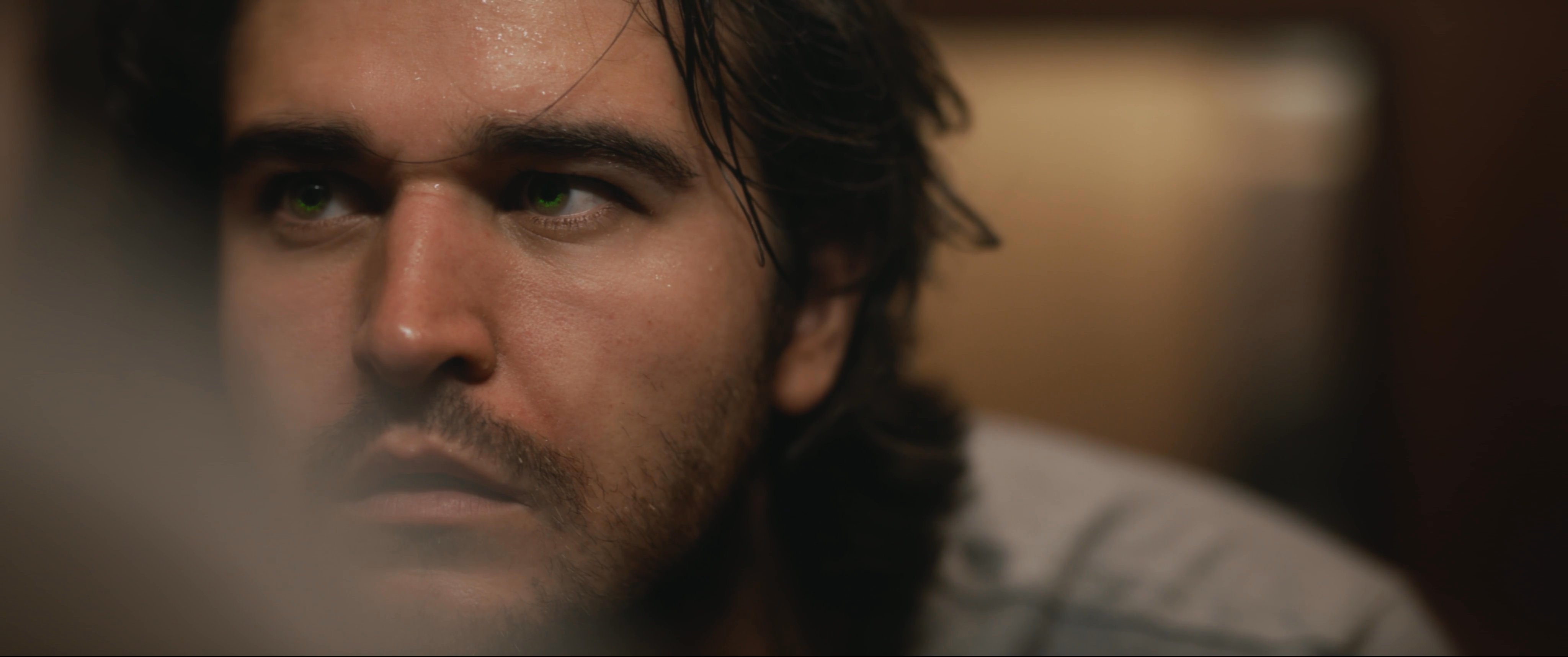 A man observes his reflection in a bathroom mirror with a focused, introspective expression while touching his face. He transitions from checking his appearance to adjusting his clothing in a dimly lit, wood-paneled room.