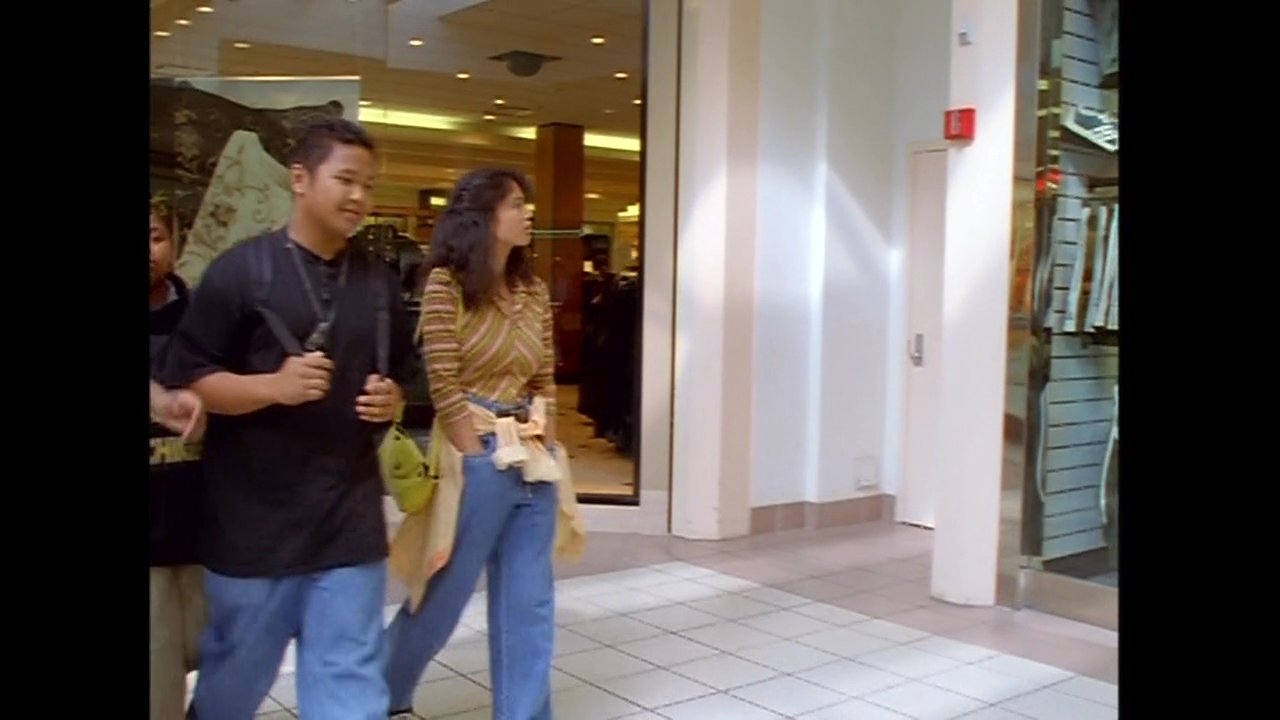 A group of young adults navigates an indoor shopping center, walking past various retail storefronts and browsing window displays.