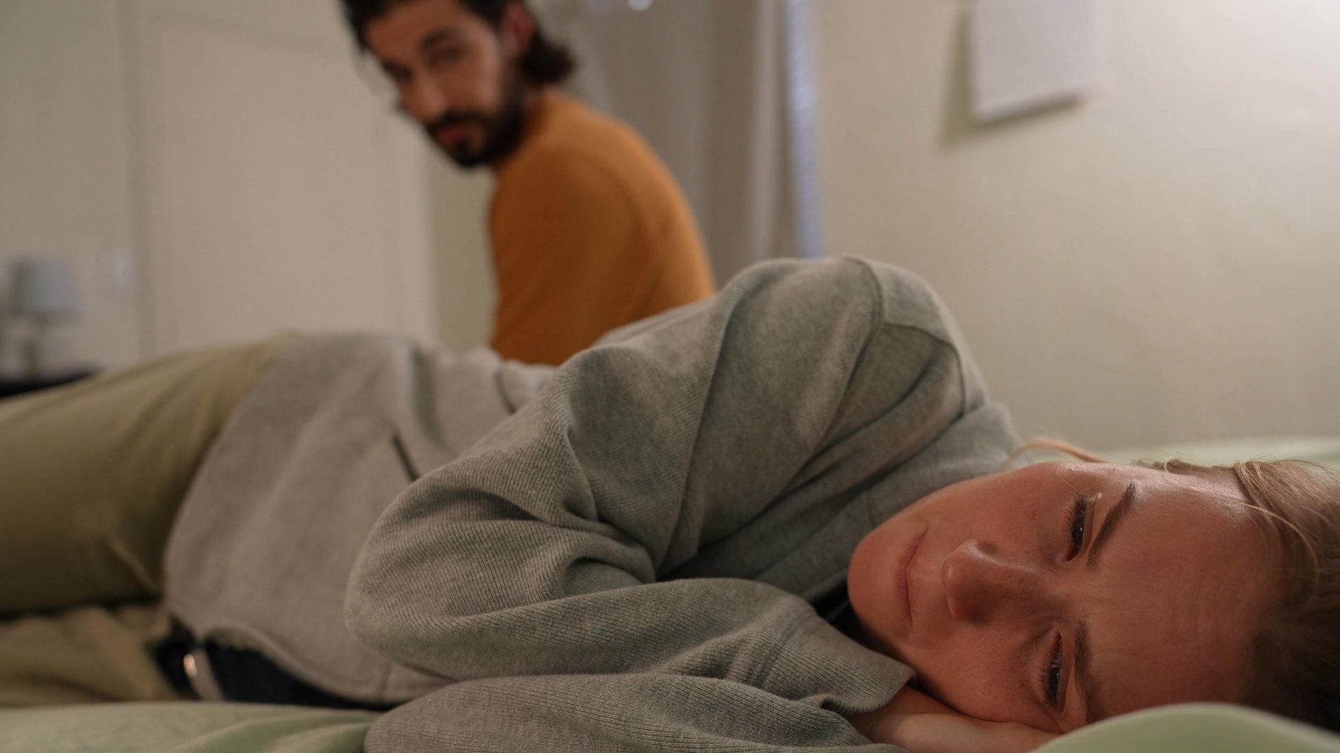 A man sits beside a woman resting on a bed as they share a quiet, contemplative moment together in a bedroom.