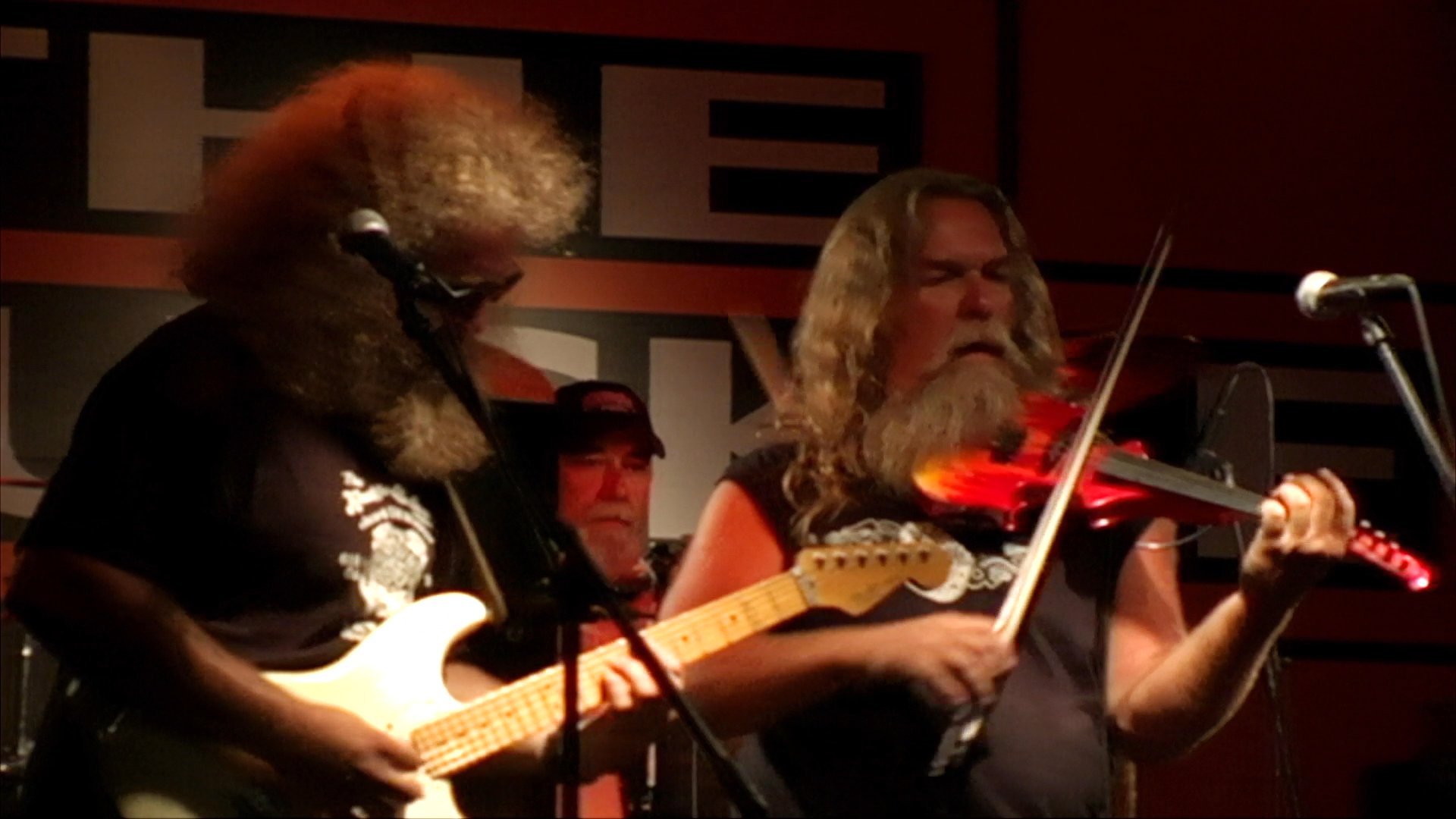 A band performs a live set on a concert stage featuring guitar and violin players. The scene captures the high-energy musical atmosphere and transitions to a brief, vintage-style outdoor gathering.
