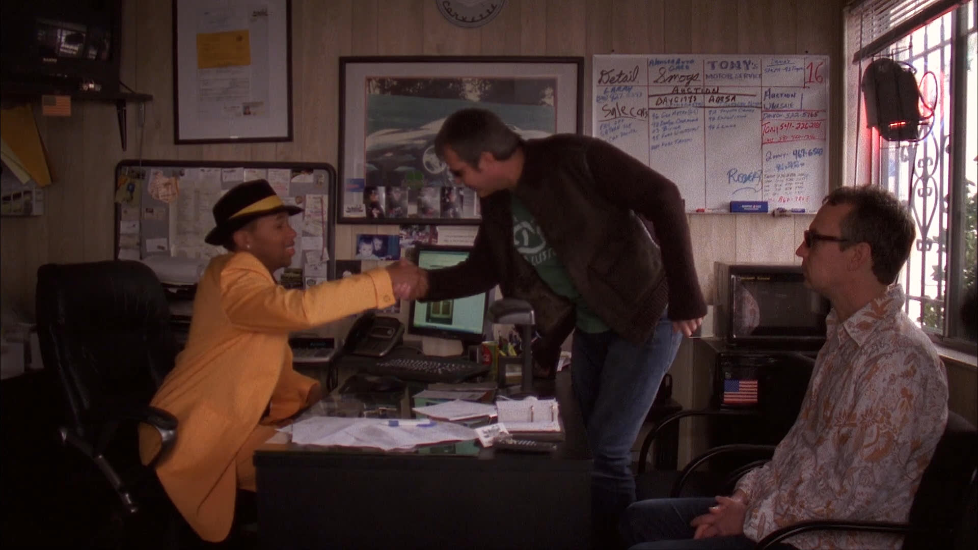 Two men arrive at a used car dealership and negotiate a vehicle purchase with the salesman before departing for a test drive in a classic car.