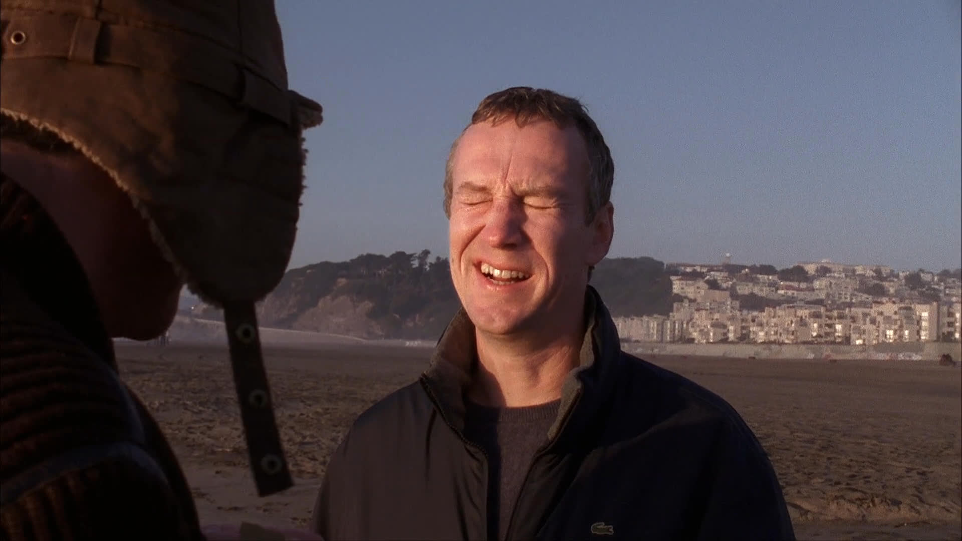 Two individuals engage in a lighthearted conversation on a beach while one manipulates a dog puppet equipped with a camera.
