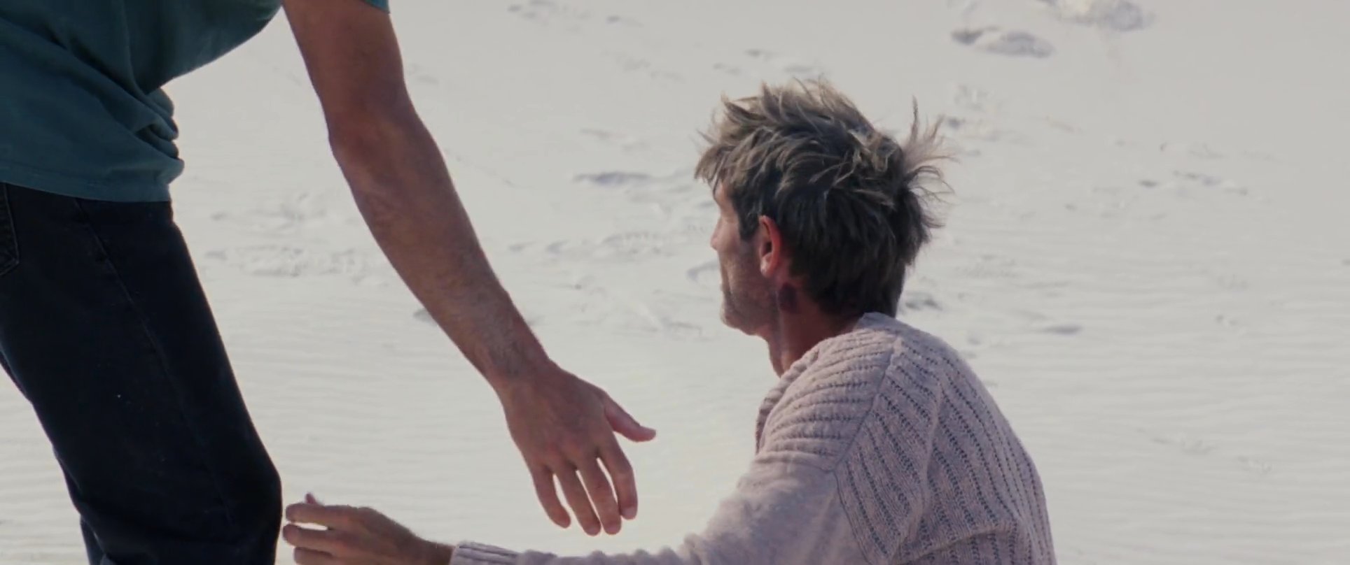One man reaches out to assist another up from the sand in a vast desert landscape, culminating in a supportive embrace between the two men.