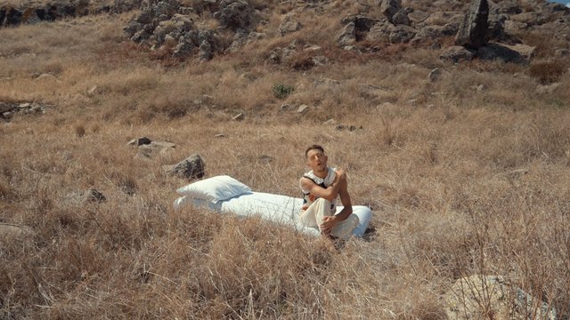 A guy is sitting on a mattress while singing in a dry field
