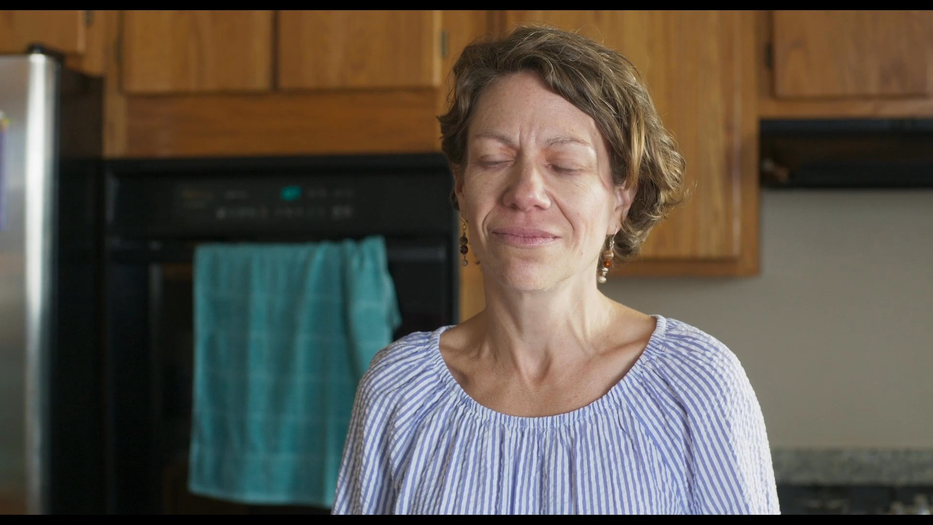 A woman stands in a kitchen and speaks while a young person sits at a table in the background. The interaction reflects a domestic exchange within a residential setting.