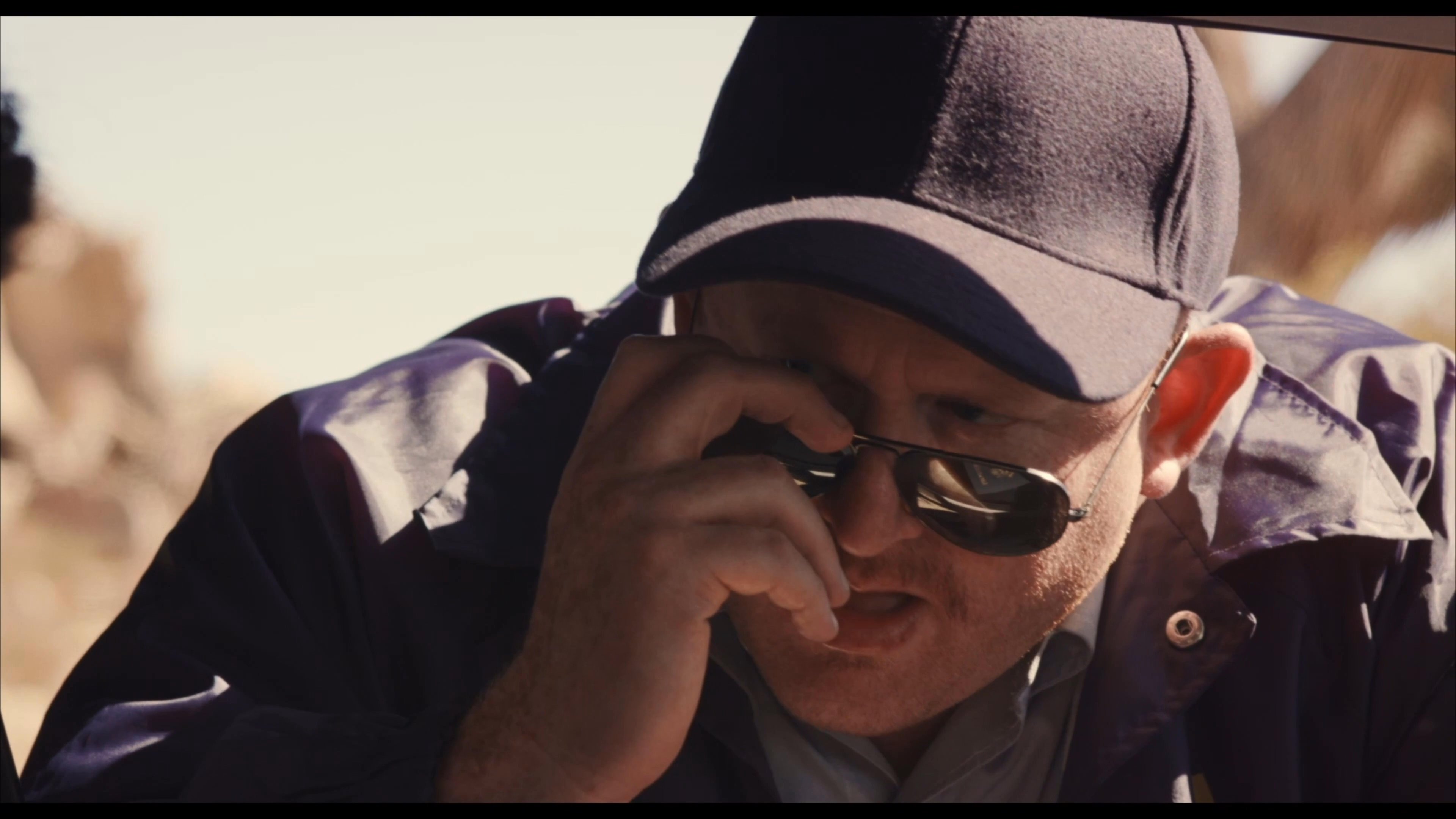 An individual in a cap and sunglasses directs occupants inside a vehicle parked in a desert environment. The scene captures a moment of focused instruction and interpersonal tension within the constrained space of the car.
