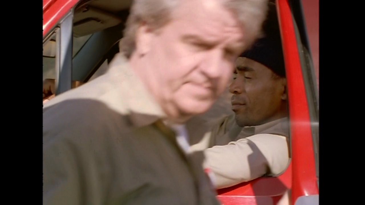 Two men in work uniforms conclude a conversation beside a red pickup truck before one drives away into a dry, mountainous area. The interaction captures a brief professional exchange in an isolated outdoor setting.