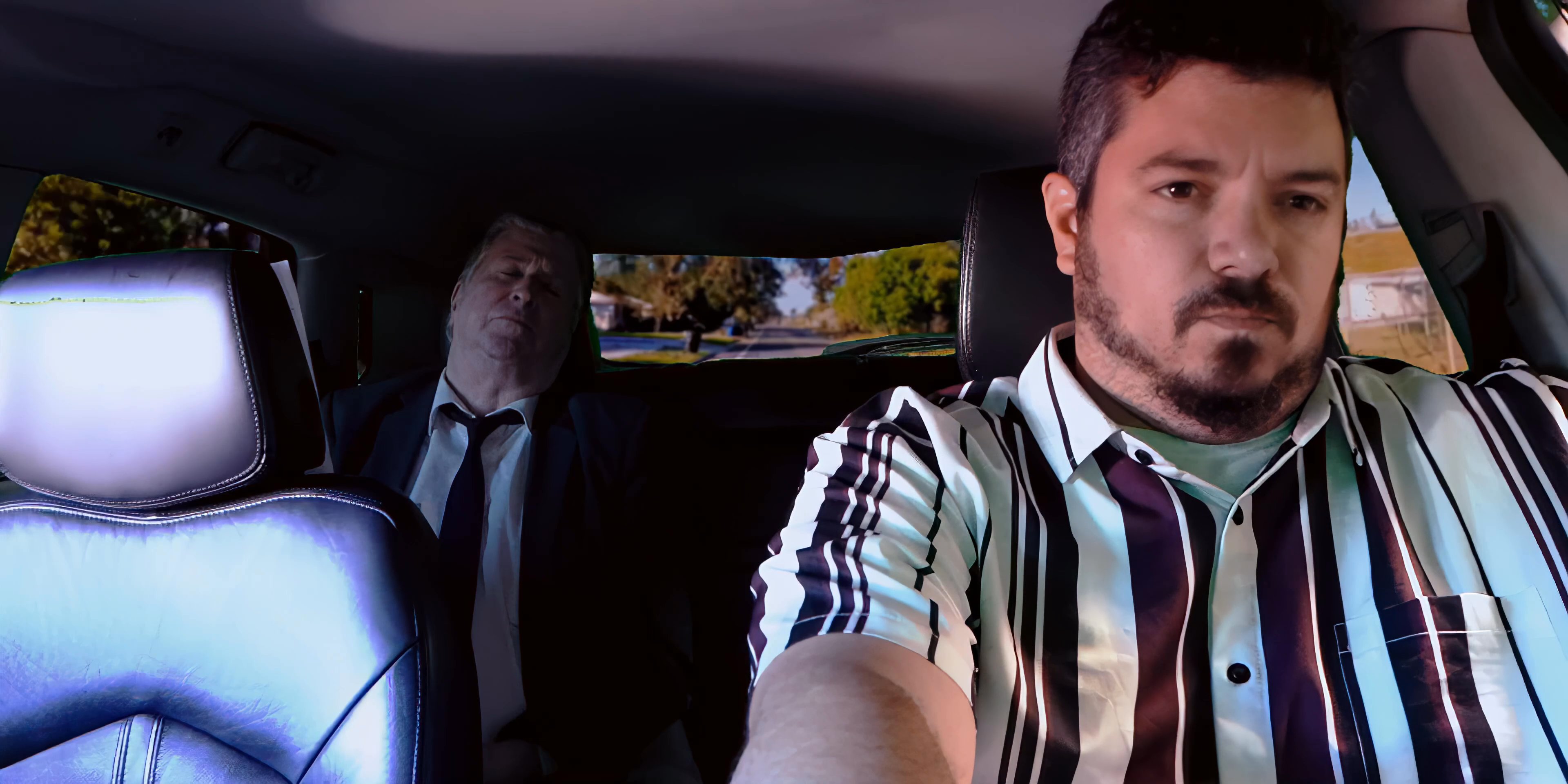 Aerial perspectives of a coastal city transition to a man driving a vehicle with an sleeping passenger in the backseat. The camera captures the commute through residential streets as the sun illuminates the afternoon landscape.
